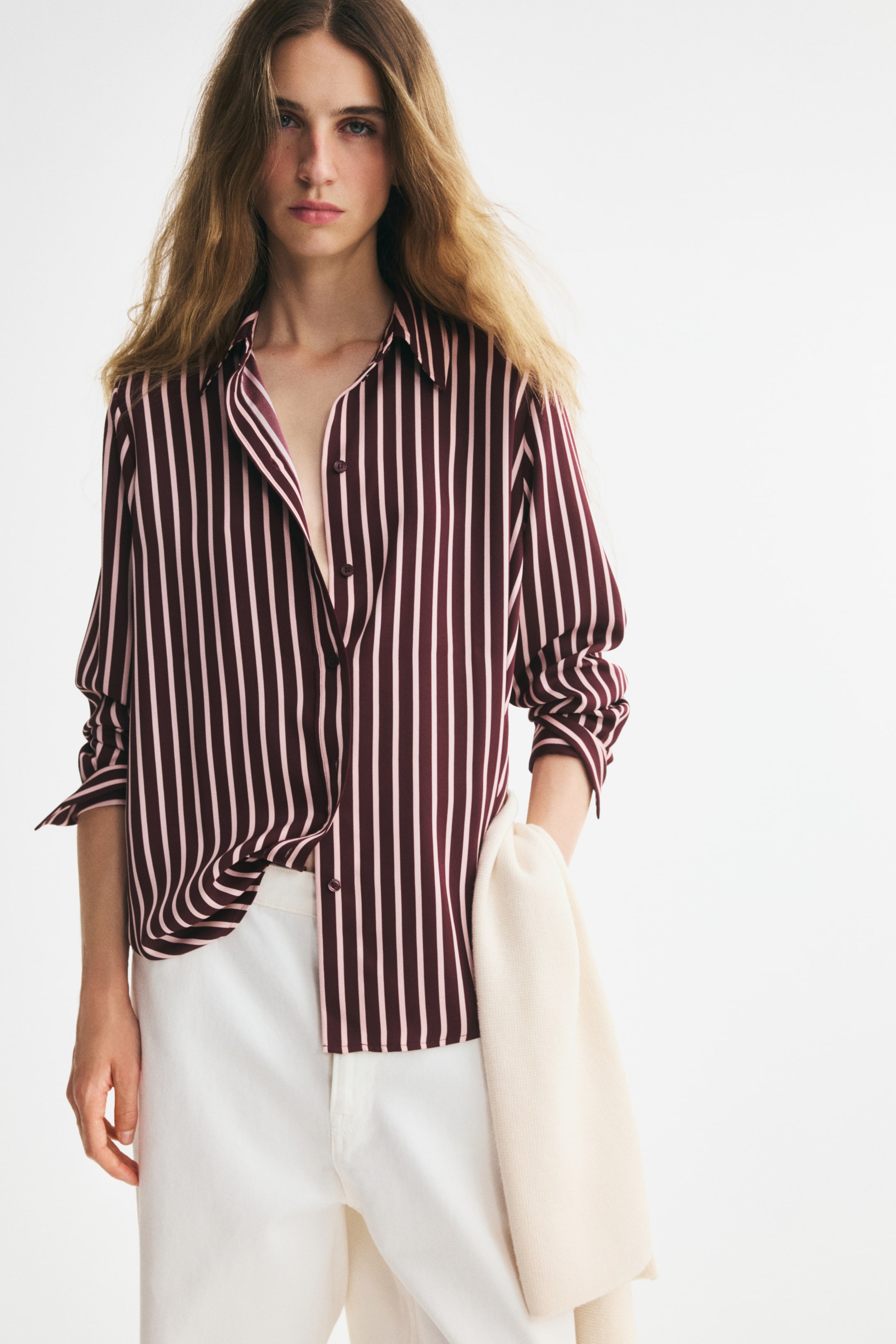 View larger image: A long-sleeved collared shirt in woven fabric is shown from the front, featuring a burgundy and light pink vertical stripe pattern. The shirt has a button-down front, is worn partially open at the neck, and has a rounded hem, partially tucked into white trousers.