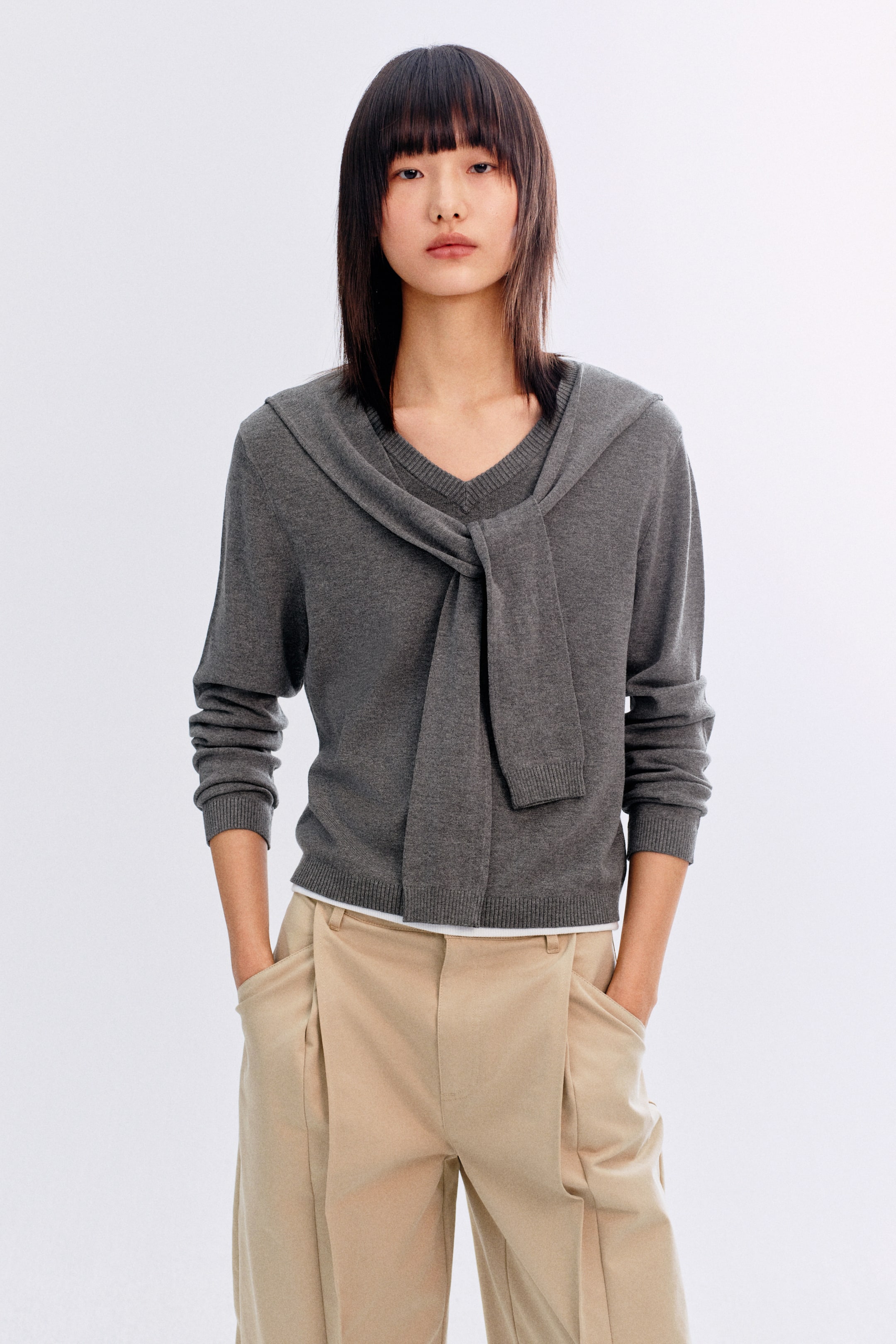 이미지 확대 보기: 모델이 착용하고 있는 제품은 V넥 라인의 짙은 회색 점퍼로, 긴 소매와 목선, 소매, 밑단에 골지 디테일이 있습니다. 어깨에 묶인 점퍼처럼 보이도록 디자인된 넓은 골지 트림 디테일이 목선을 감싸고 앞쪽에서 매듭지어져 있습니다. 이 점퍼는 흰색 이너셔츠 위에 레이어링되어 있으며, 밝은 갈색의 주름 바지와 스타일링되어 있습니다.