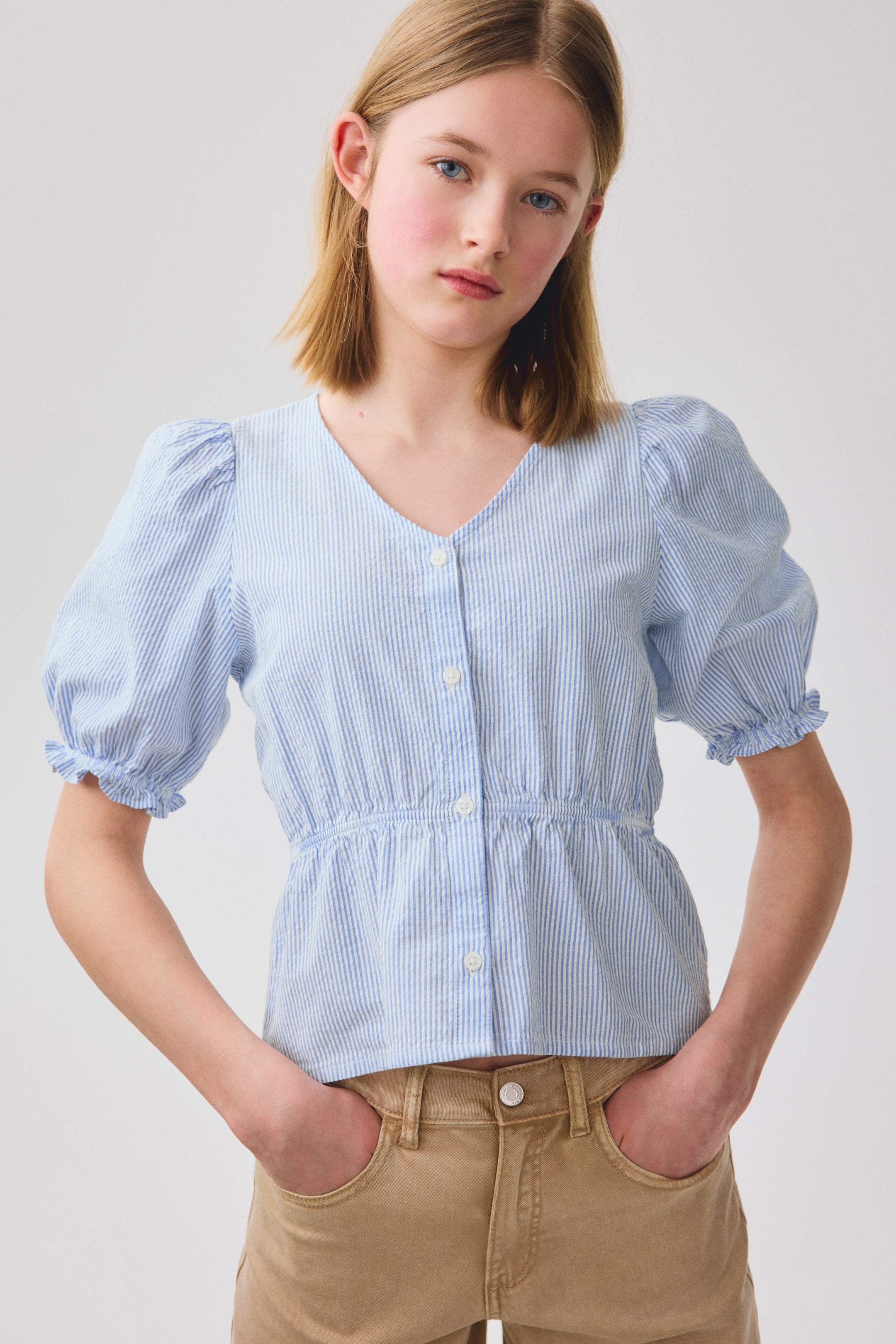 View larger image: An airy blue and white striped cotton seersucker blouse is shown from the front. It features a V-shaped neckline, buttons down the front, and narrow elastic at the waist, along with short puff sleeves with elasticated, frill-trimmed cuffs.