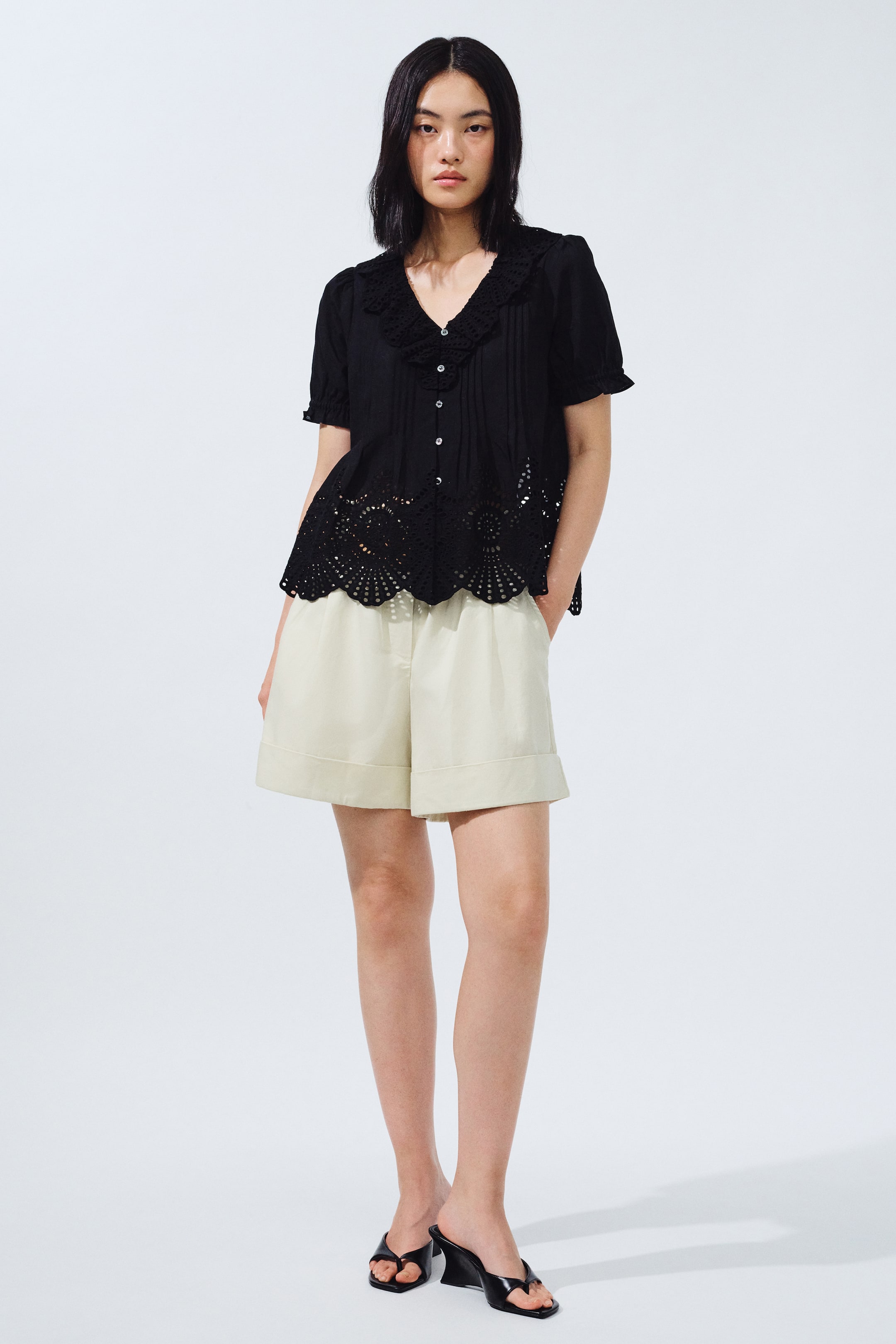 View larger image: The model is donning a black short-sleeve top featuring a ruffled V-neck, pintucks, and eyelet cutwork detailing at the hem. She pairs it with light beige cuffed shorts, casually placing her hands in the pockets, and completes the look with black heeled thong sandals.