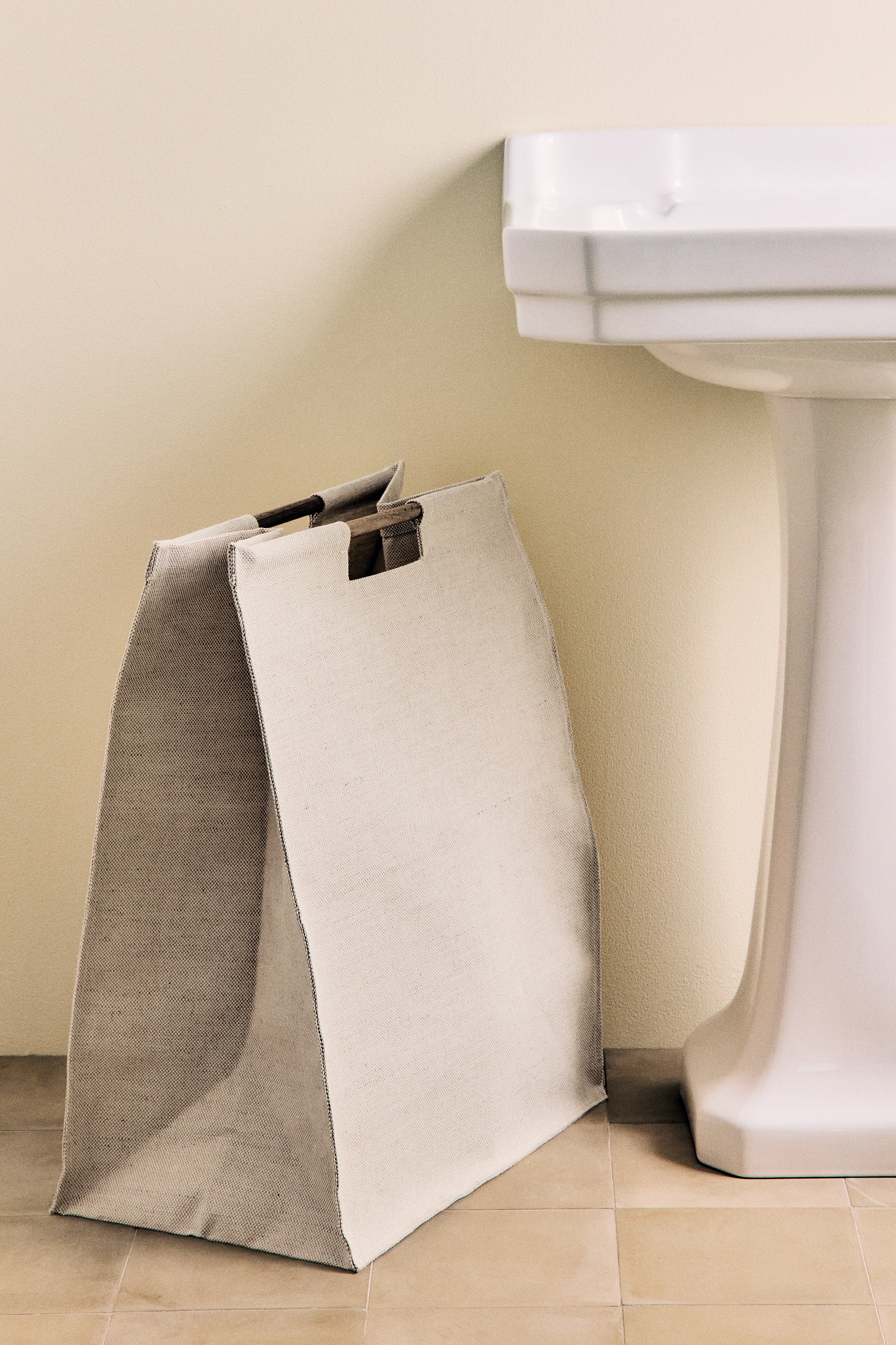 The rectangular hamper is presenting a front view, constructed from a light beige woven fabric with a visible texture. It is featuring two horizontal wooden bar handles integrated into its top edge.