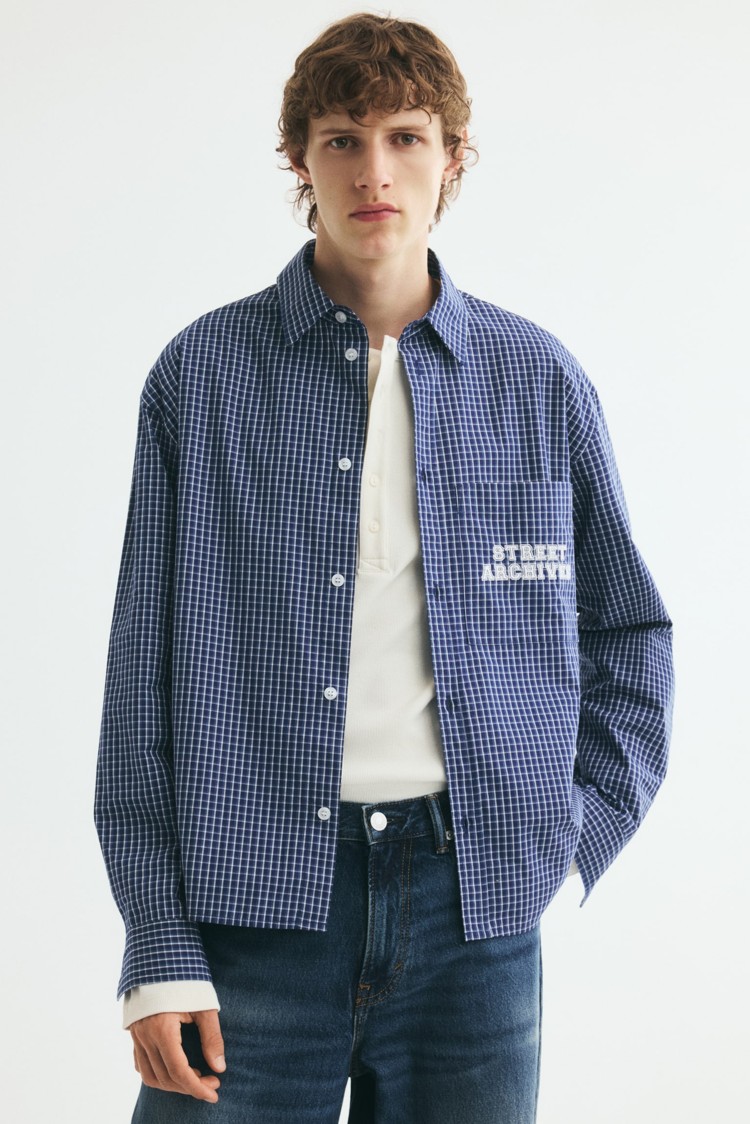 A blue and white checkered cotton poplin shirt is being worn open by the model, layered over a cream-colored knit top and dark blue jeans. The relaxed-fit shirt features a classic collar, dropped shoulders, and a patch pocket on the chest with white printed text. - 1