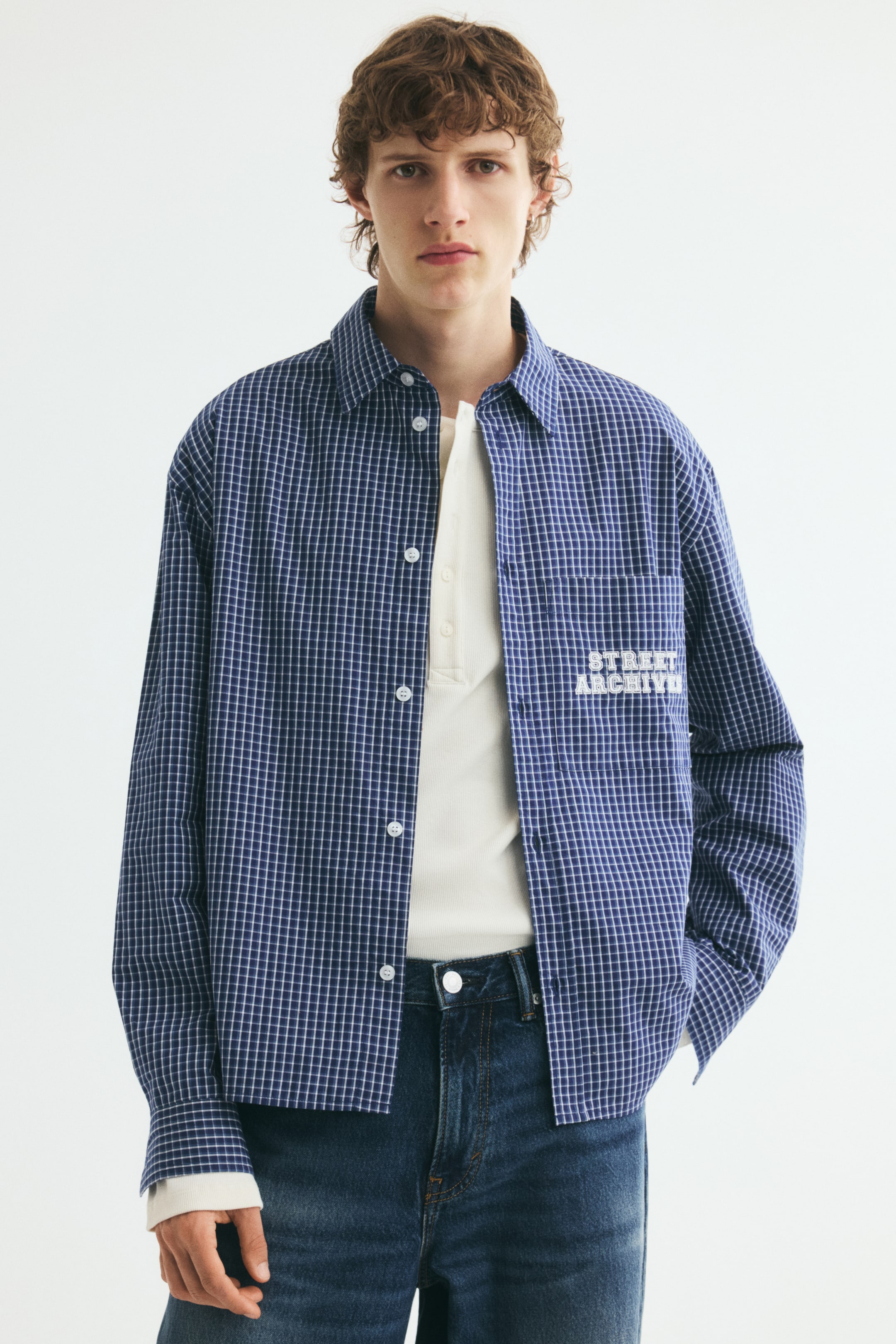 View larger image: A blue and white checkered cotton poplin shirt is worn open by the model, layered over a cream-coloured knit top and dark blue jeans. The relaxed-fit shirt features a classic collar, dropped shoulders, and a patch pocket on the chest with white printed text.