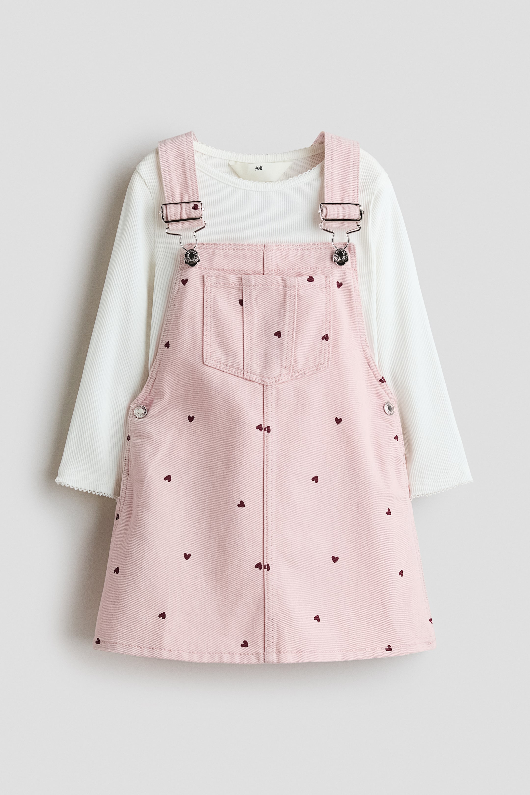 View larger image: A pink overall dress, featuring a bib front with a patch pocket, silver-toned buckles, and scattered dark red heart prints, is showcased from the front. It is styled over a white ribbed long-sleeved top with a scalloped neckline and cuffs.
