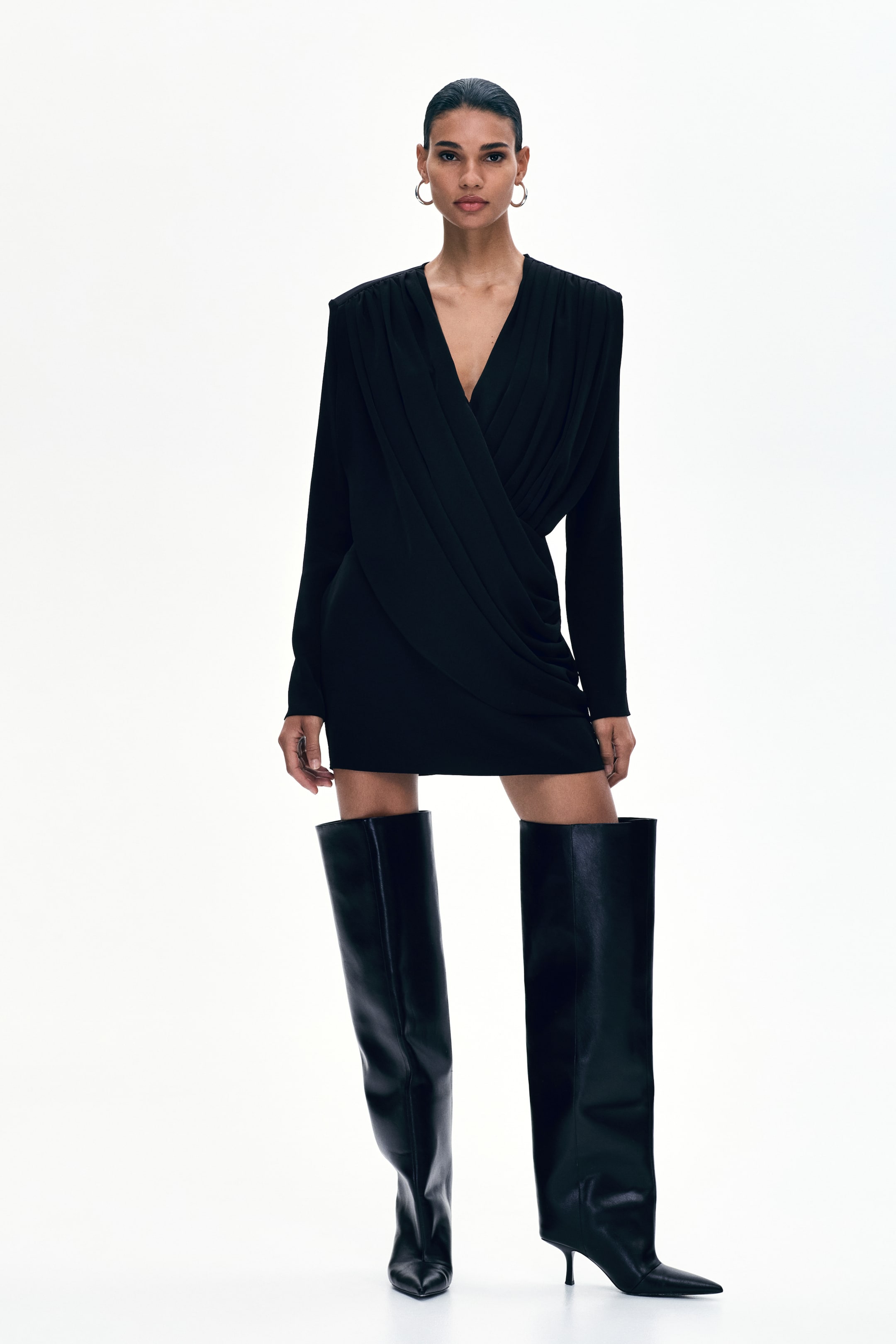 View larger image: The model is showcasing a short black dress, highlighted by a draped wrapover front, a deep V-neckline, and long sleeves with shoulder gathers that shape a defined silhouette. The dress boasts a fitted skirt, complemented by the model's tall black boots.