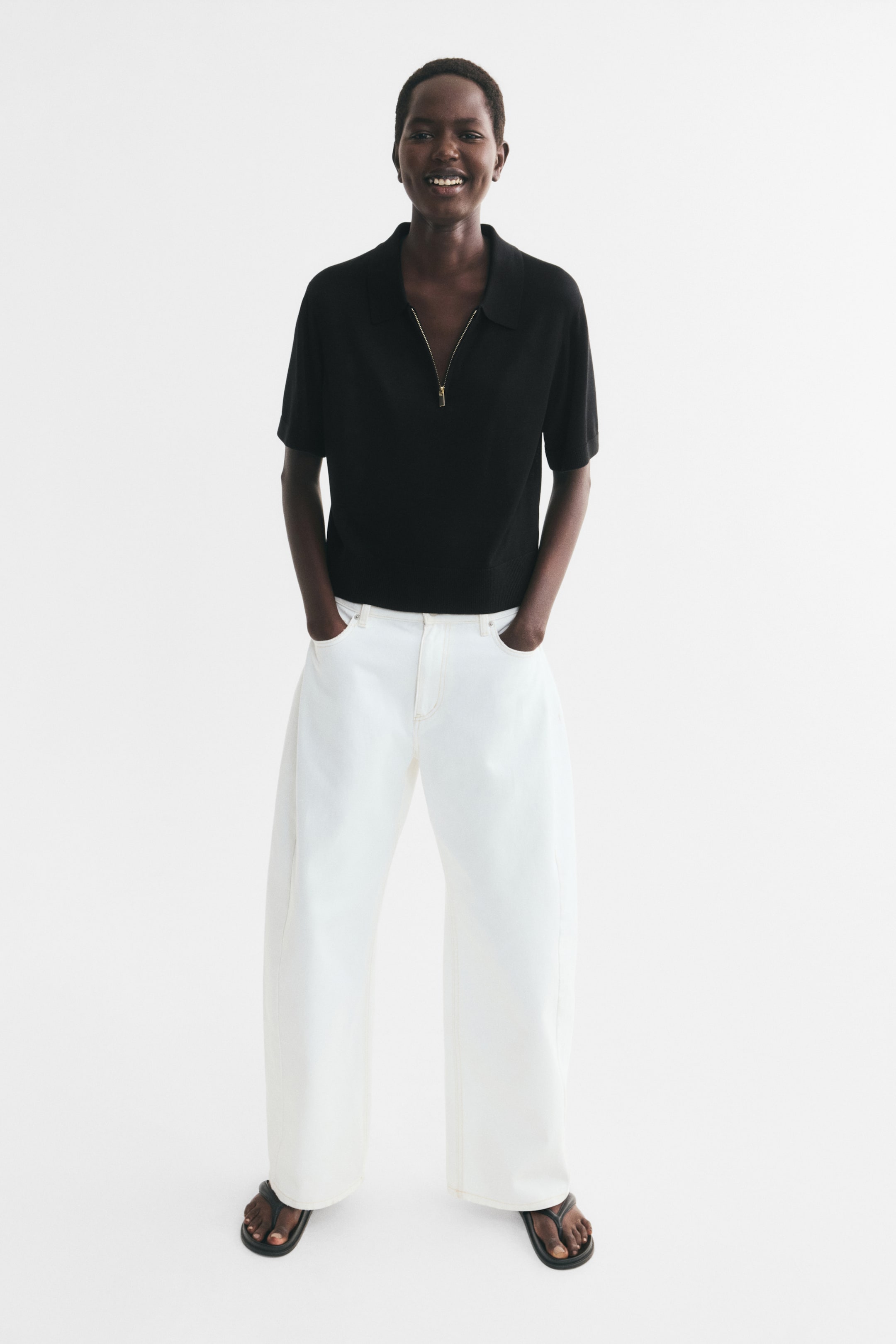 View larger image: The model is wearing a black, short-sleeved fitted polo shirt with a fine-knit texture. It features a collar, a visible gold-toned quarter zip at the neckline, and ribbed cuffs and hem.