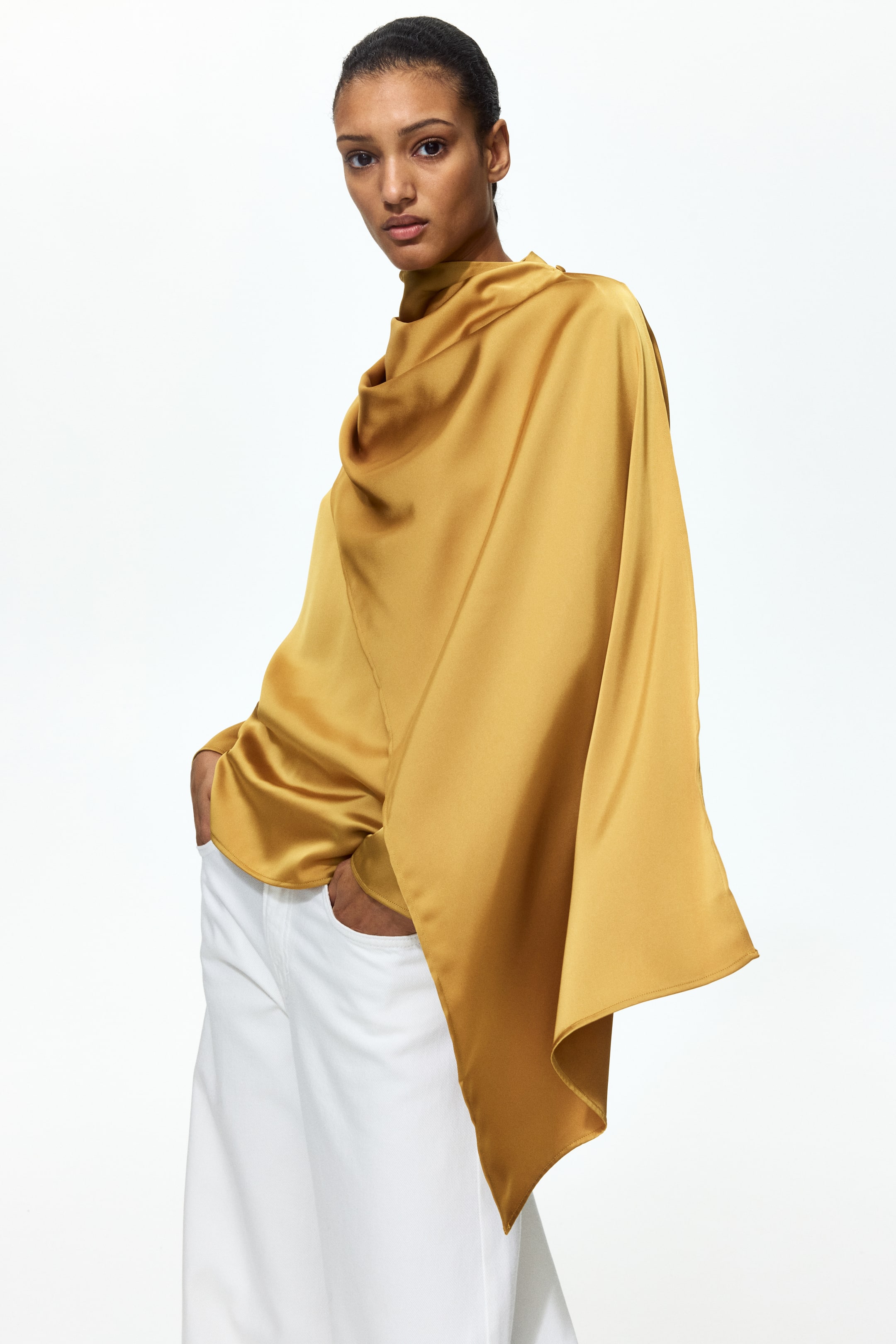 Größeres Bild anzeigen: Eine glänzende Satinbluse mit Stehkragen und einem über die Vorderseite drapierten Schaldetail wird von dem Model getragen. Lange Ärmel sind sichtbar, und die Hand des Models ruht in der Tasche einer weißen Hose.