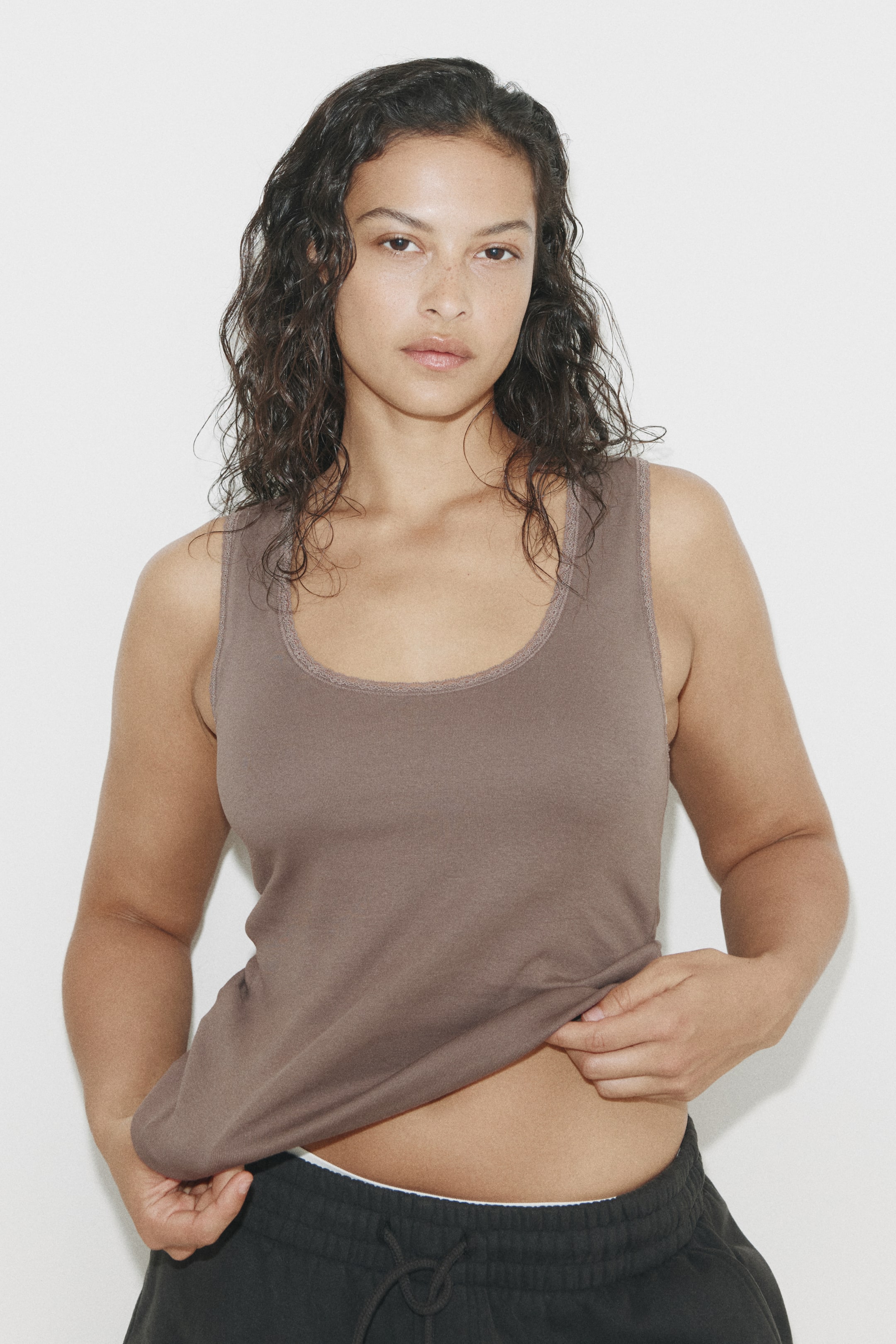 View larger image: A brown tank top featuring a scoop neckline with delicate, slightly textured trim along the neck and armholes. The fitted top is slightly pulled up at the hem, revealing its smooth knit fabric.