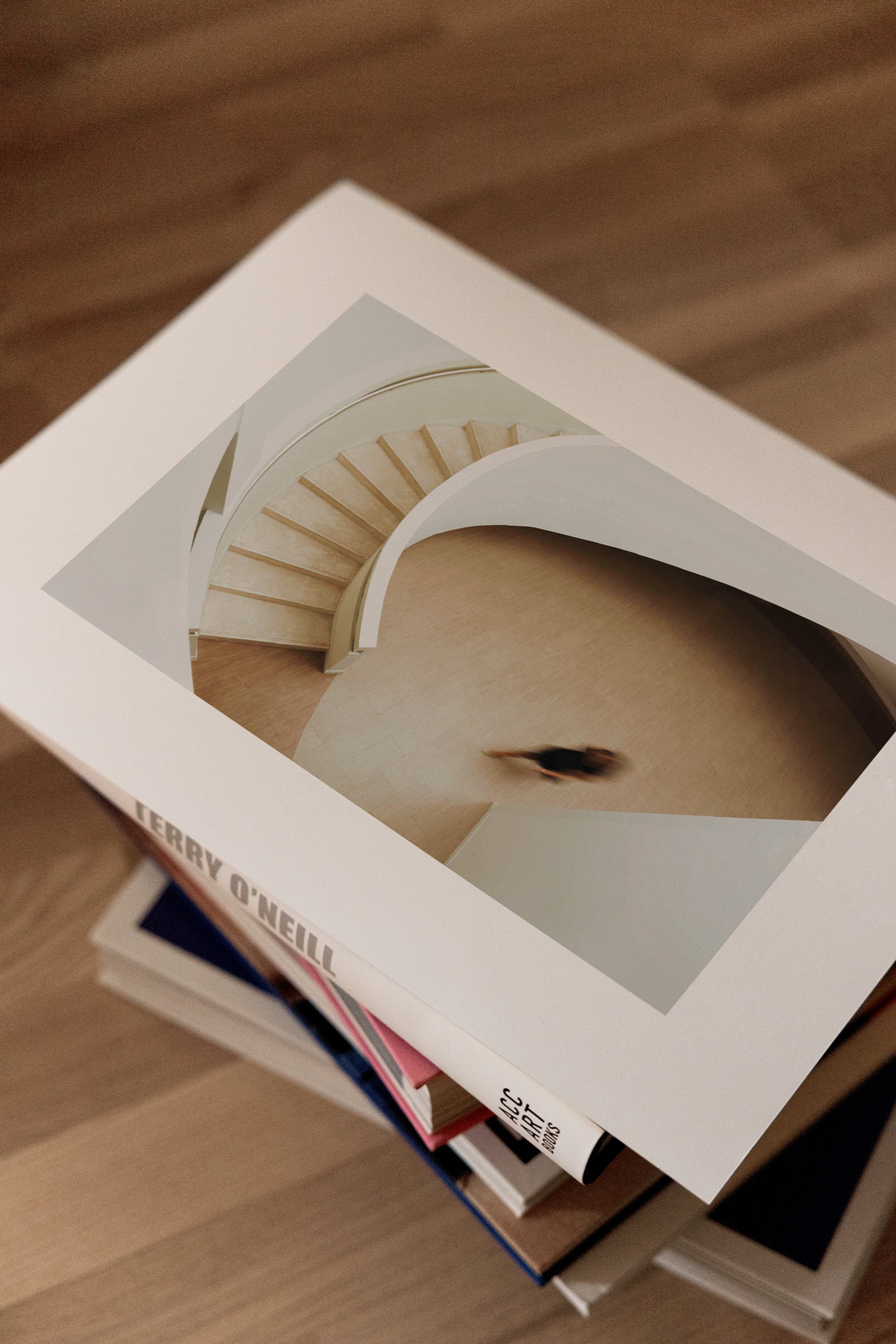 Se større billede: En hvid hardcoverbog med titlen "Terry O'Neill" ligger diagonalt på en stak bøger og viser forsiden med et minimalistisk spiraltrappemotiv og en sløret figur. Bogens ryg viser titlen og forlagslogoet "ACC ART BOOKS" i et rent, sort skriftbillede.