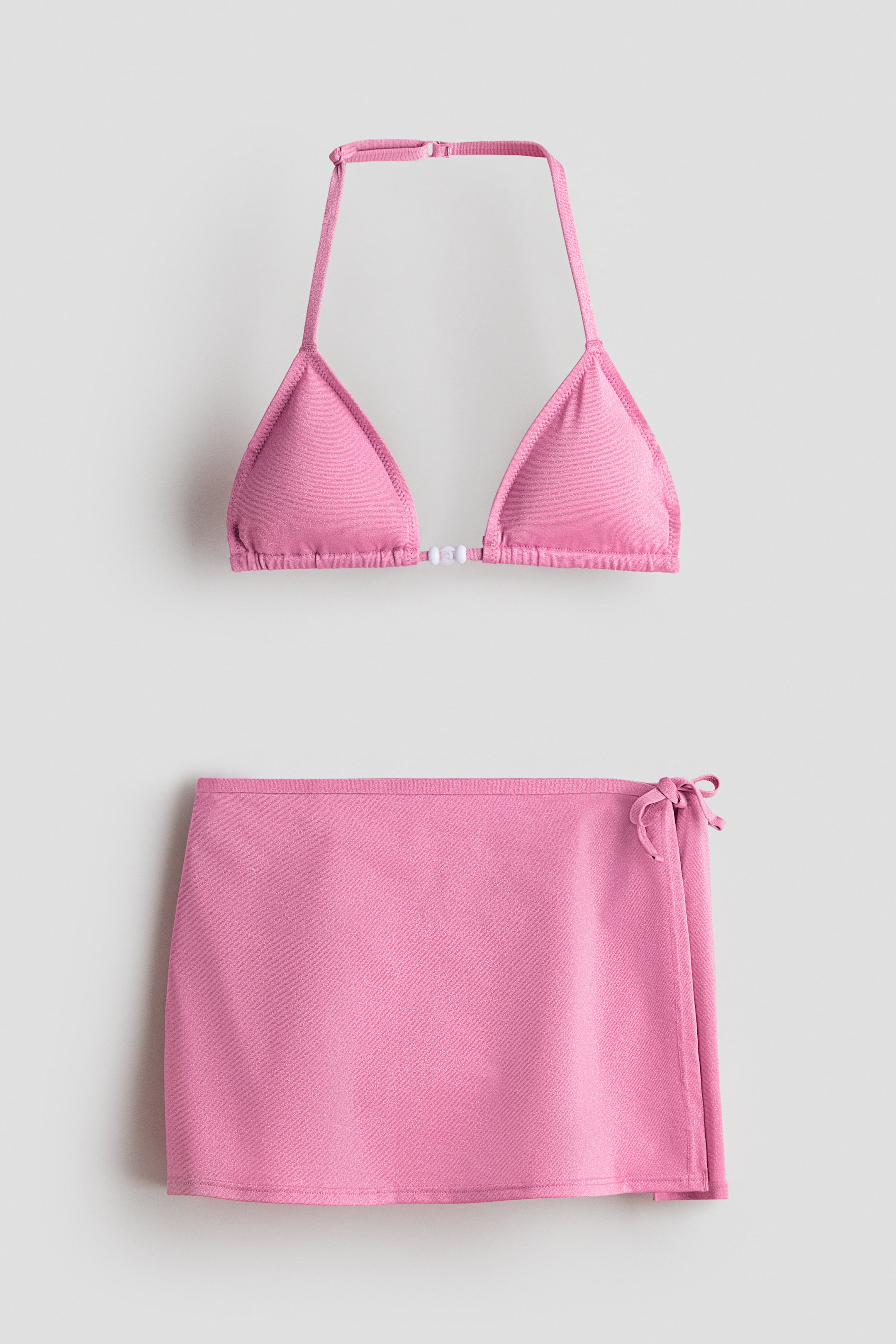 Se større billede: Et lyserødt glitrende bikinisæt med en trekantet haltertop, der har små hvide perler, som forbinder skålene. Den matchende front wrap mini-nederdel har en bindelukning på højre side.