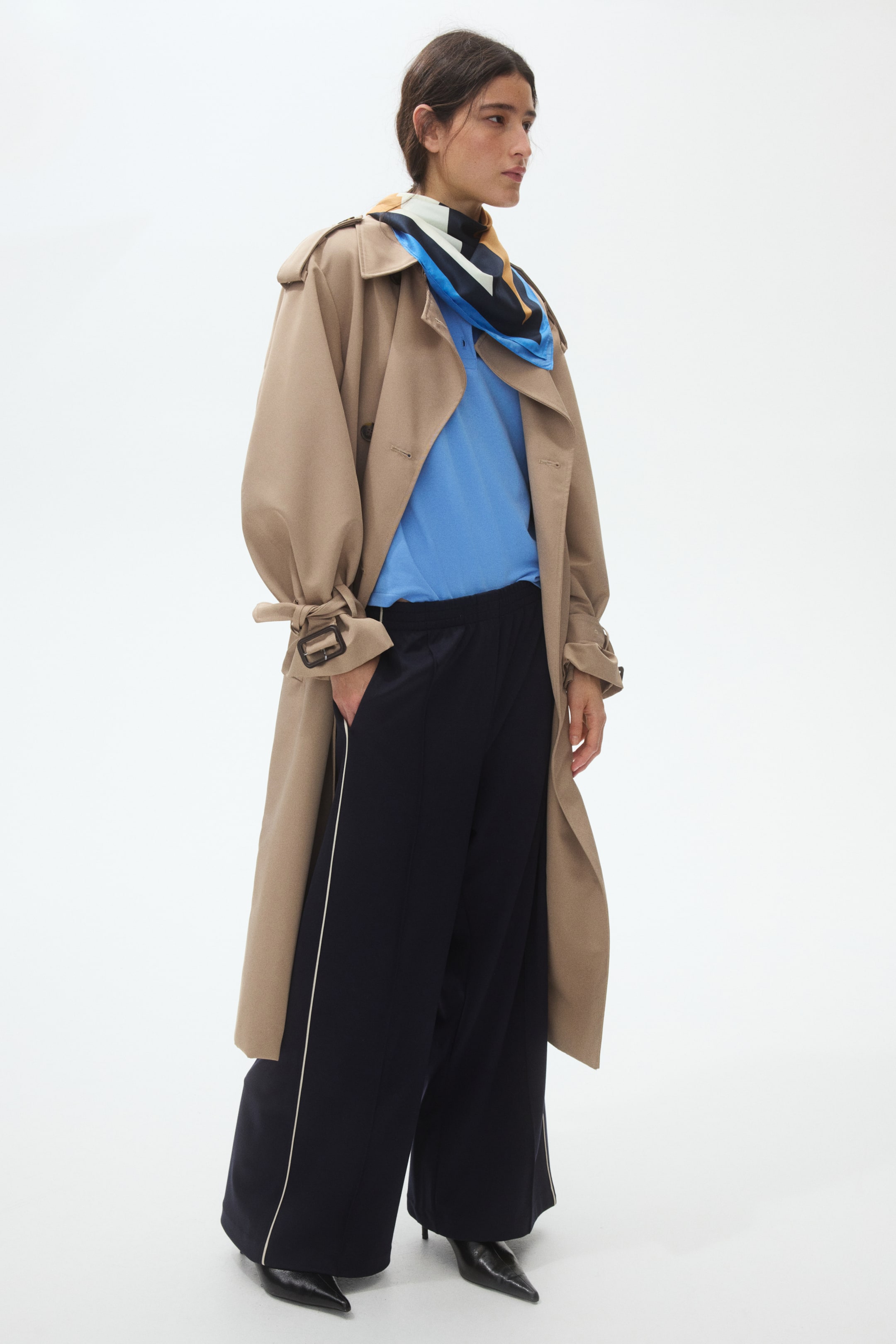 View larger image: A tan trench coat is showcased from a slight side angle, worn open over a light blue polo shirt, black wide-leg trousers with white side stripes, and a multi-coloured geometric scarf around the neck. The trench coat features gathered sleeves with buckle straps, and the wide-leg trousers flow over black pointed-toe shoes.
