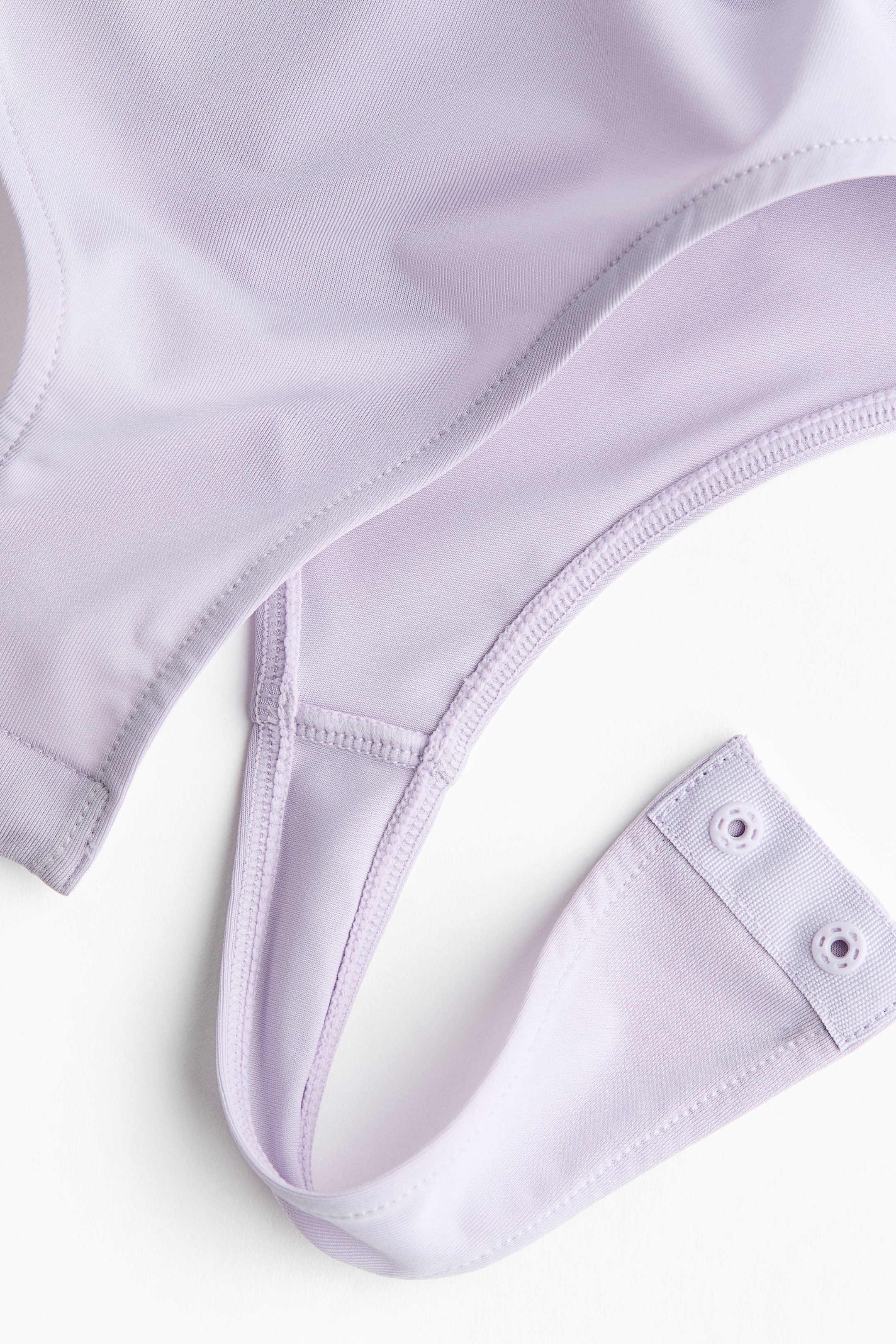 View larger image: A lavender thong bodysuit features smooth fabric with subtle ribbing. The thong-style bottom is visible, ending in a tab with two light purple snap closures.