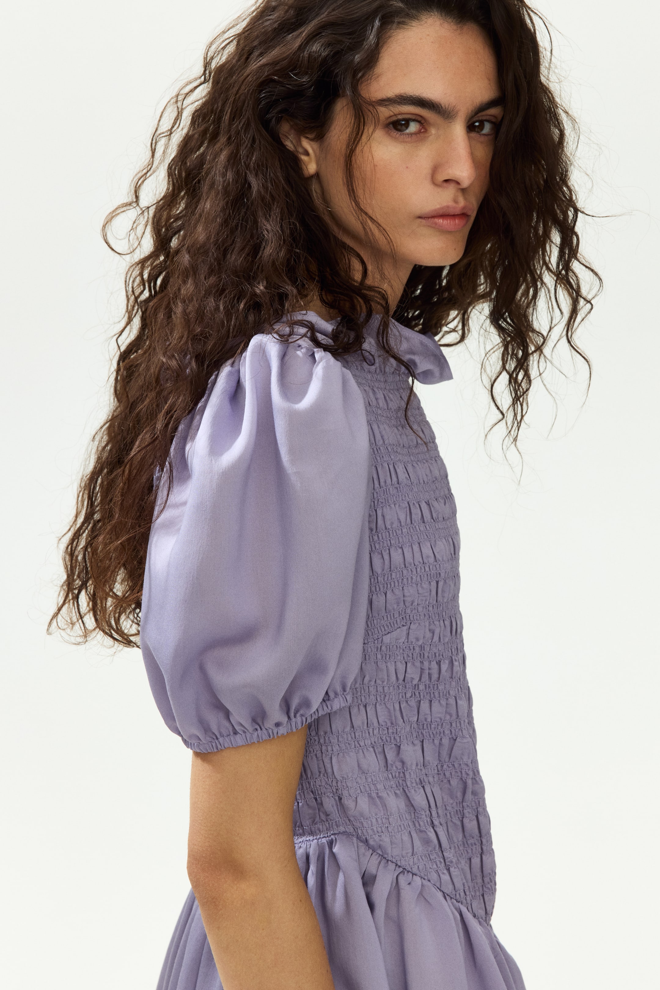 View larger image: A soft lavender dress is highlighting a smocked bodice, short puff sleeves, and a gathered peplum. The sleeves are gathered at the shoulders and hems, while the bodice is showcasing extensive horizontal smocking.
