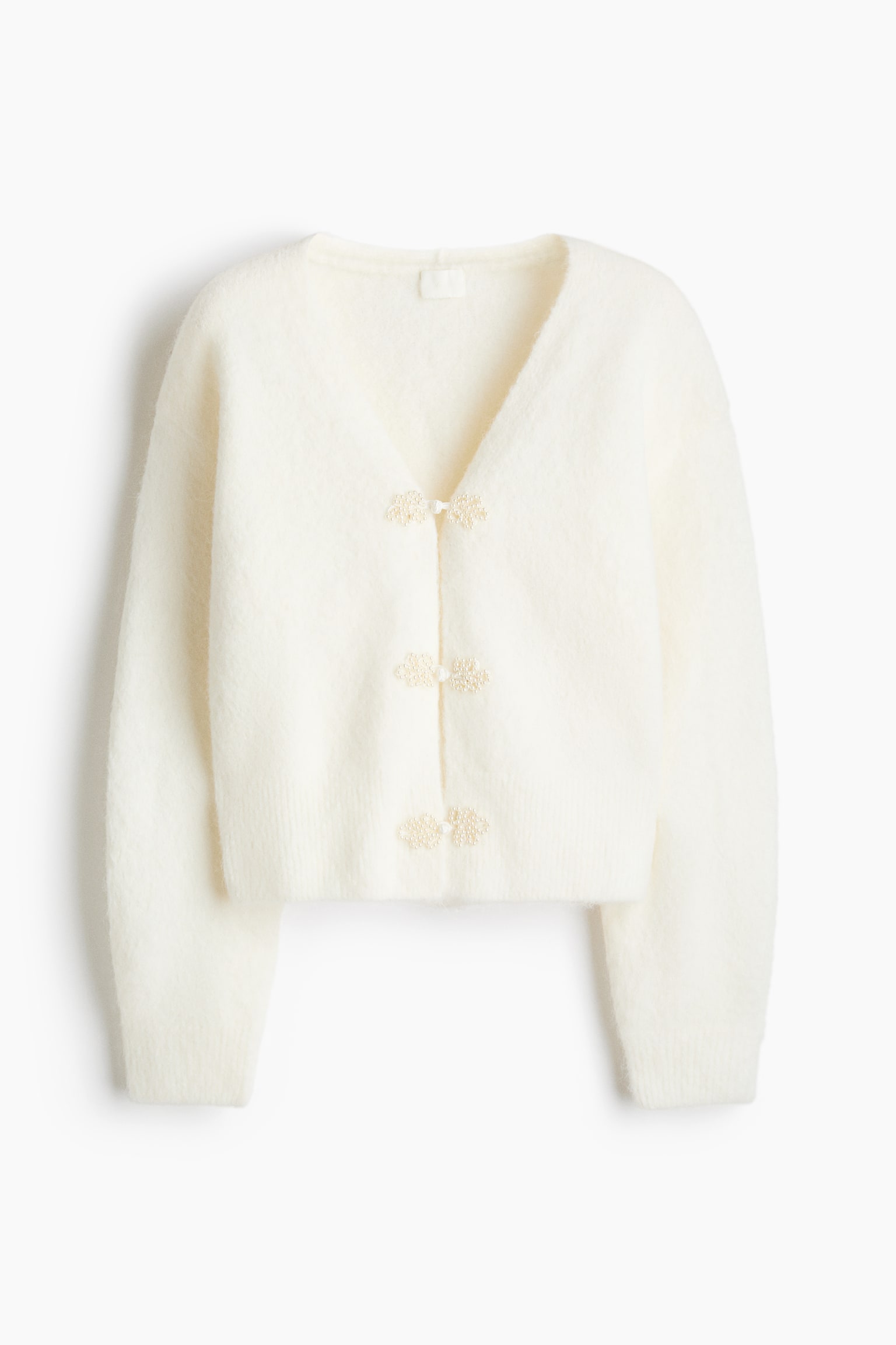 Un cardigan lavorato a maglia color crema, morbido e peloso, Ã¨ ripreso frontalmente e mette in risalto un profondo scollo a V, maniche lunghe con polsini a coste e un orlo a coste. Il davanti Ã¨ chiuso con tre alamari decorativi impreziositi da perle.