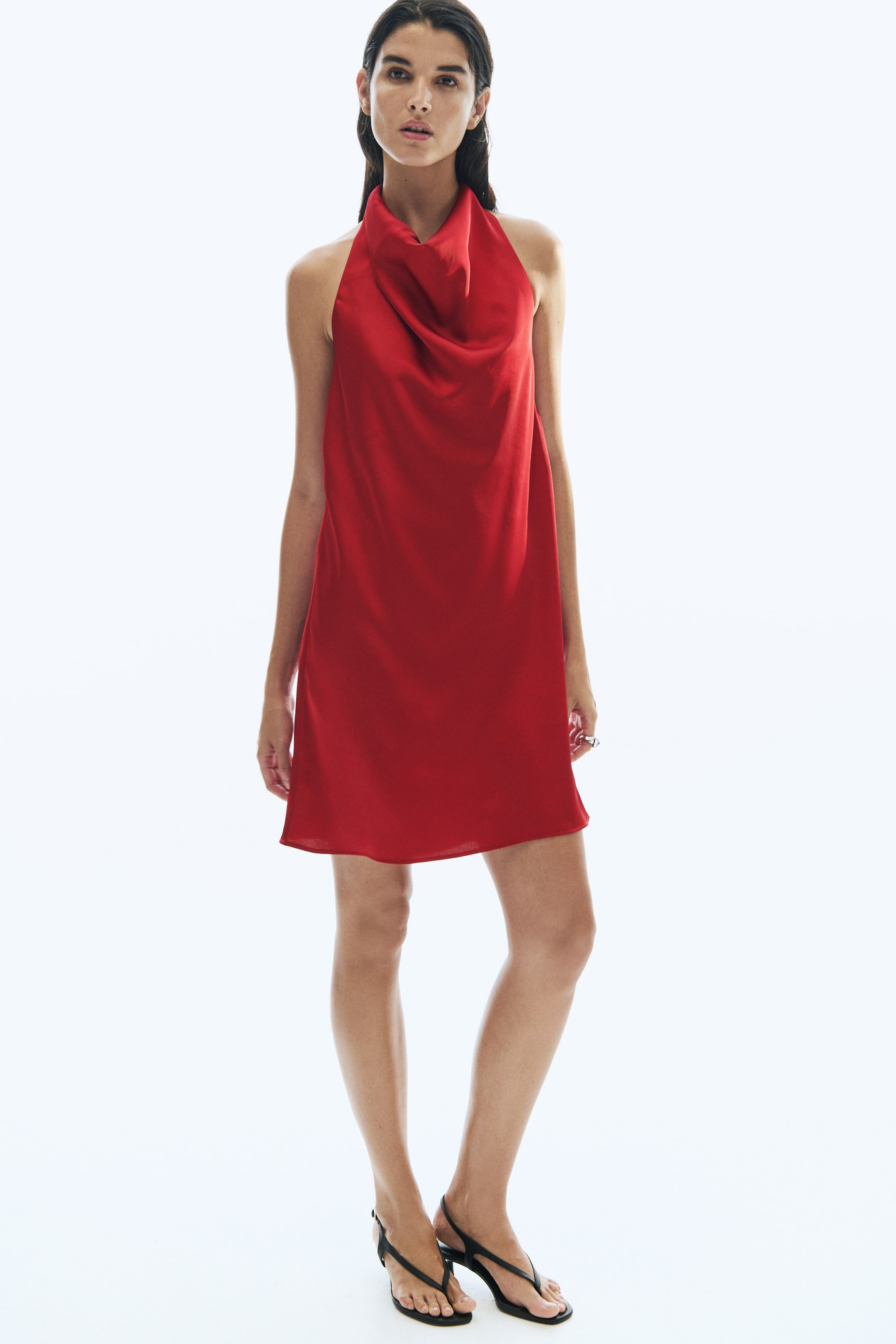 View larger image: A red, loose-fit satin mini dress featuring a draped cowl neckline is being worn by the model standing in a forward-facing pose. The dress is styled with black thong-style sandals.