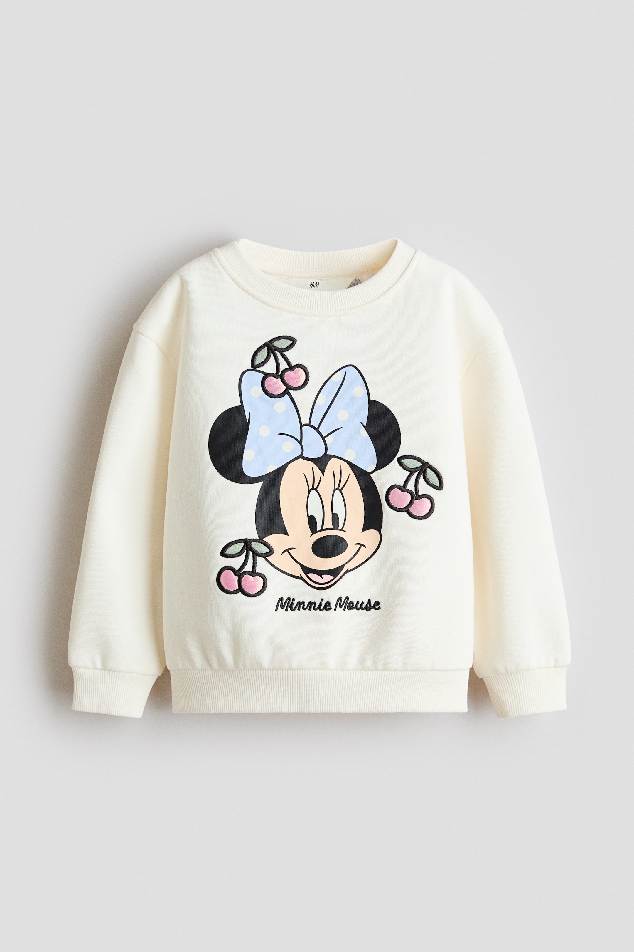 View larger image: A cream sweatshirt is showcased from the front, highlighting a Minnie Mouse print motif with cherries on its chest. It features dropped shoulders, long sleeves, and ribbed details at the neckline, cuffs, and hem.