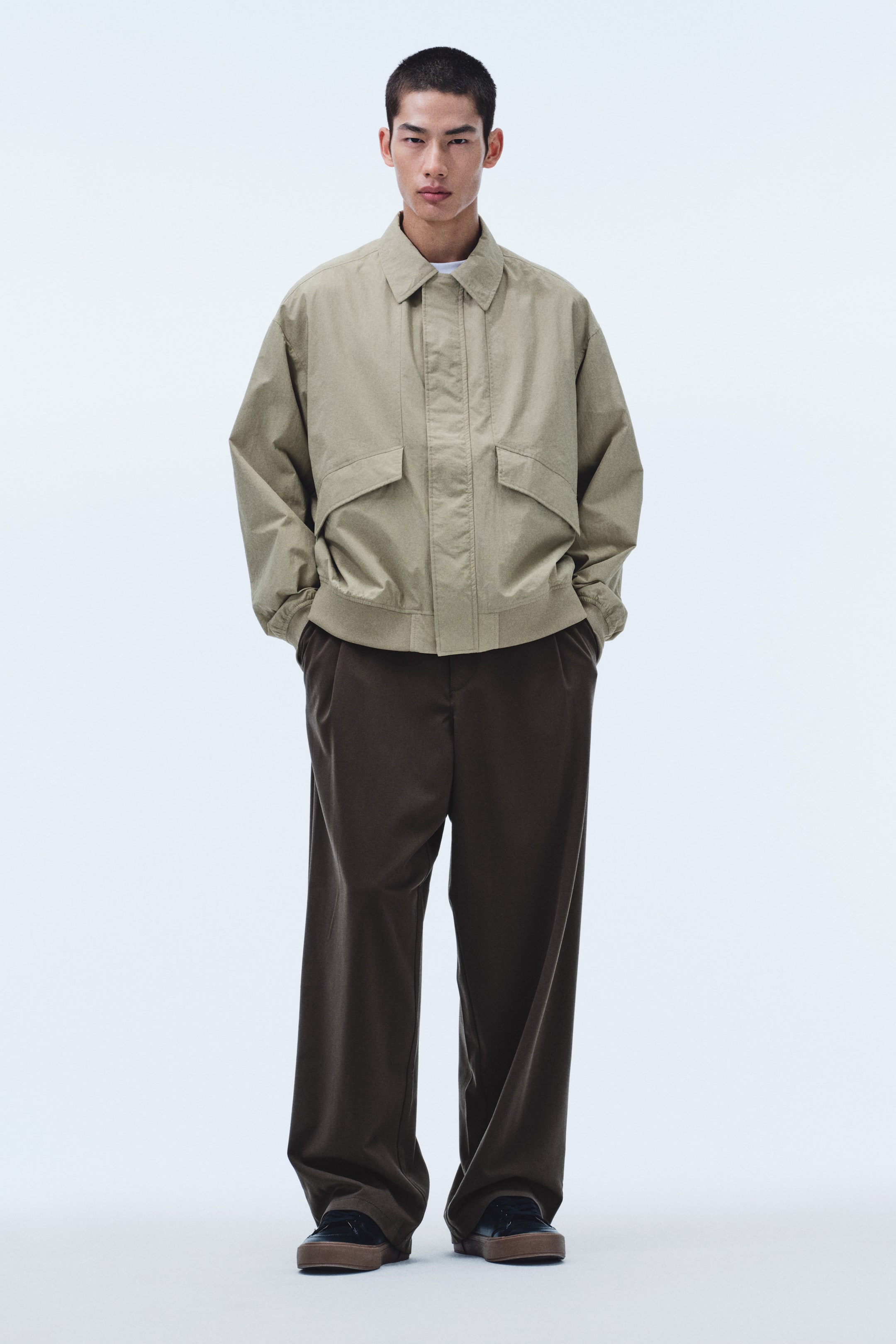 拡大画像を表示: ライトベージュのボンバージャケットは、ポインテッドカラー、比翼仕立てのフロント、フロント下部に大きめのフラップポケットが2つ、リブ仕様の袖口と裾を備え、リラックスしたフィット感を演出します。ジャケットは白いTシャツを重ねており、首元から白いTシャツがのぞいています。