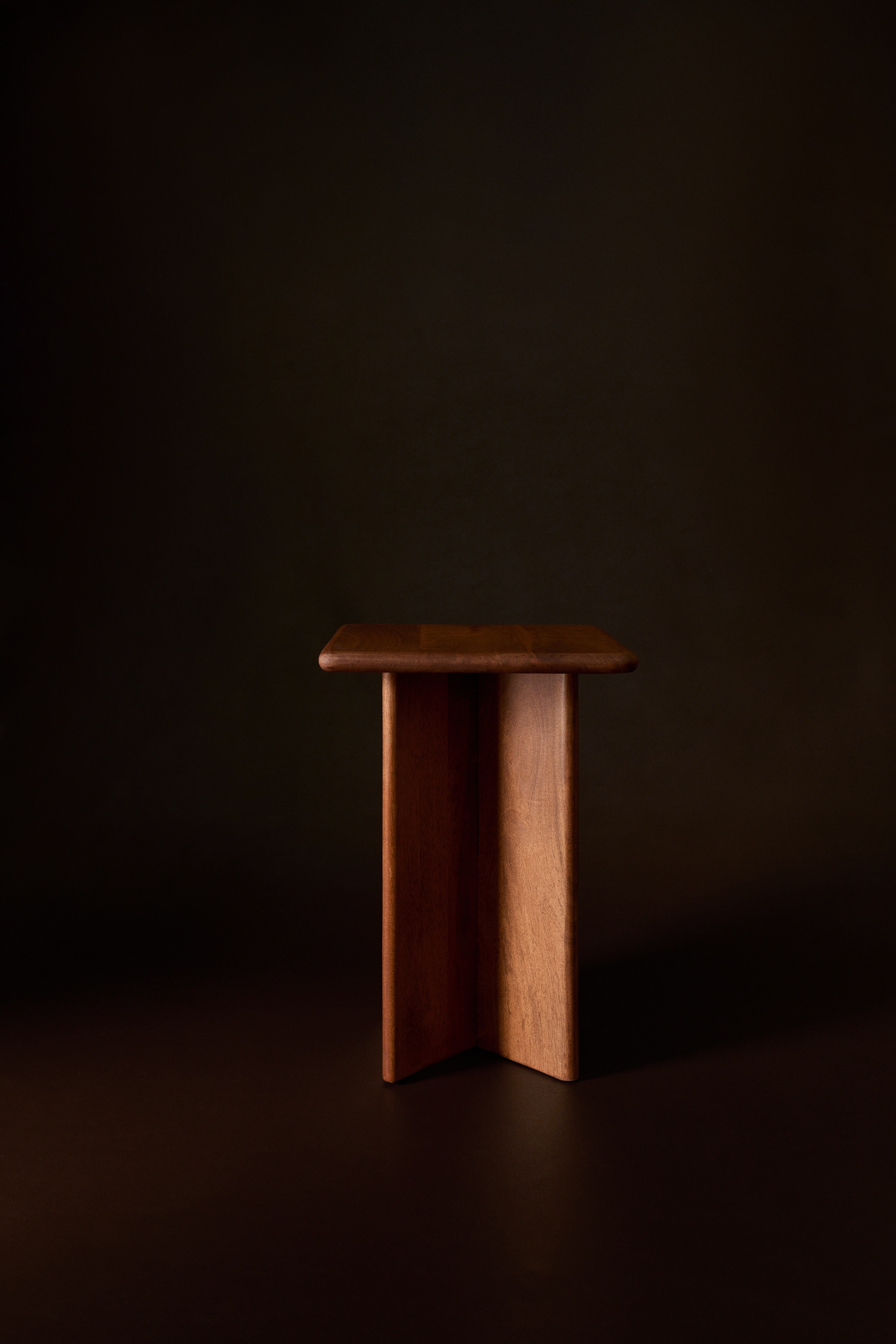 Un tavolino laterale quadrato in legno marrone medio Ã¨ mostrato da unâangolazione alta a tre quarti, con un piano spesso dai bordi e angoli arrotondati e levigati. Il tavolo Ã¨ sostenuto da una base a piedistallo a croce, composta da pannelli lignei verticali intersecati che valorizzano la venatura naturale del legno.