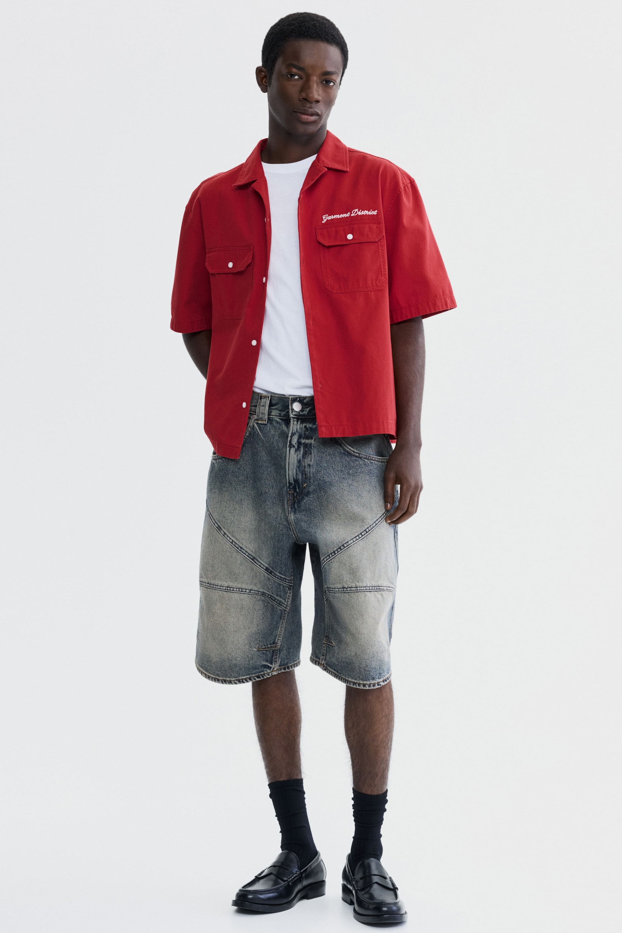 View larger image: The model is wearing a red short-sleeve collared shirt with "Garment District" embroidered on the chest, styled open over a white t-shirt. Paired with this is a pair of distressed denim shorts featuring paneled construction.