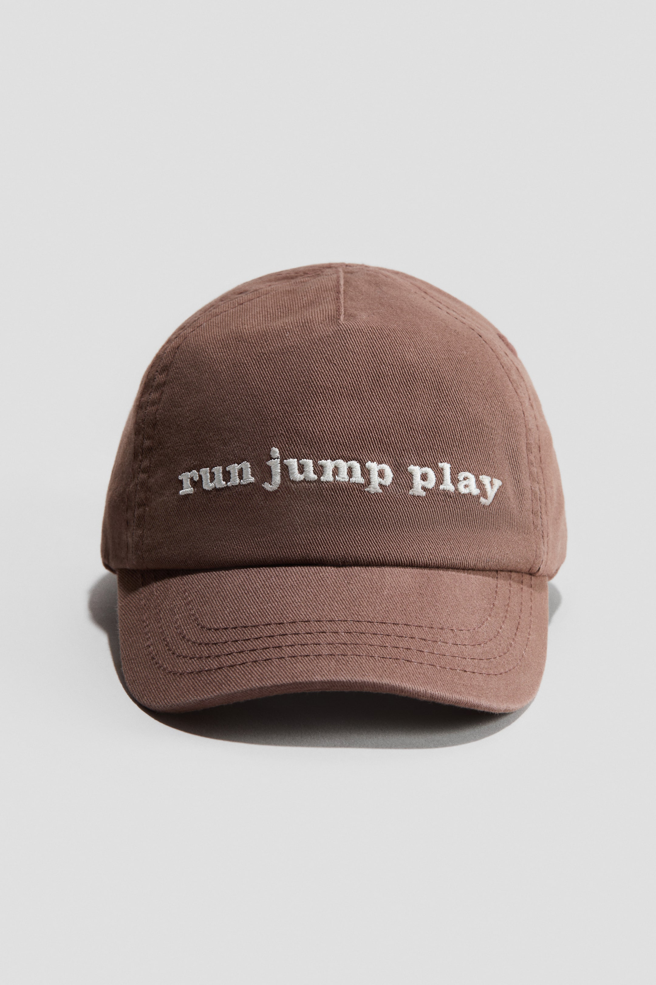 Visa större bild: En brun keps i bomullsväv är vänd framåt. Den visar vit broderad text "run jump play" över frontpanelen och har en böjd skärm med flera rader av sömmar.