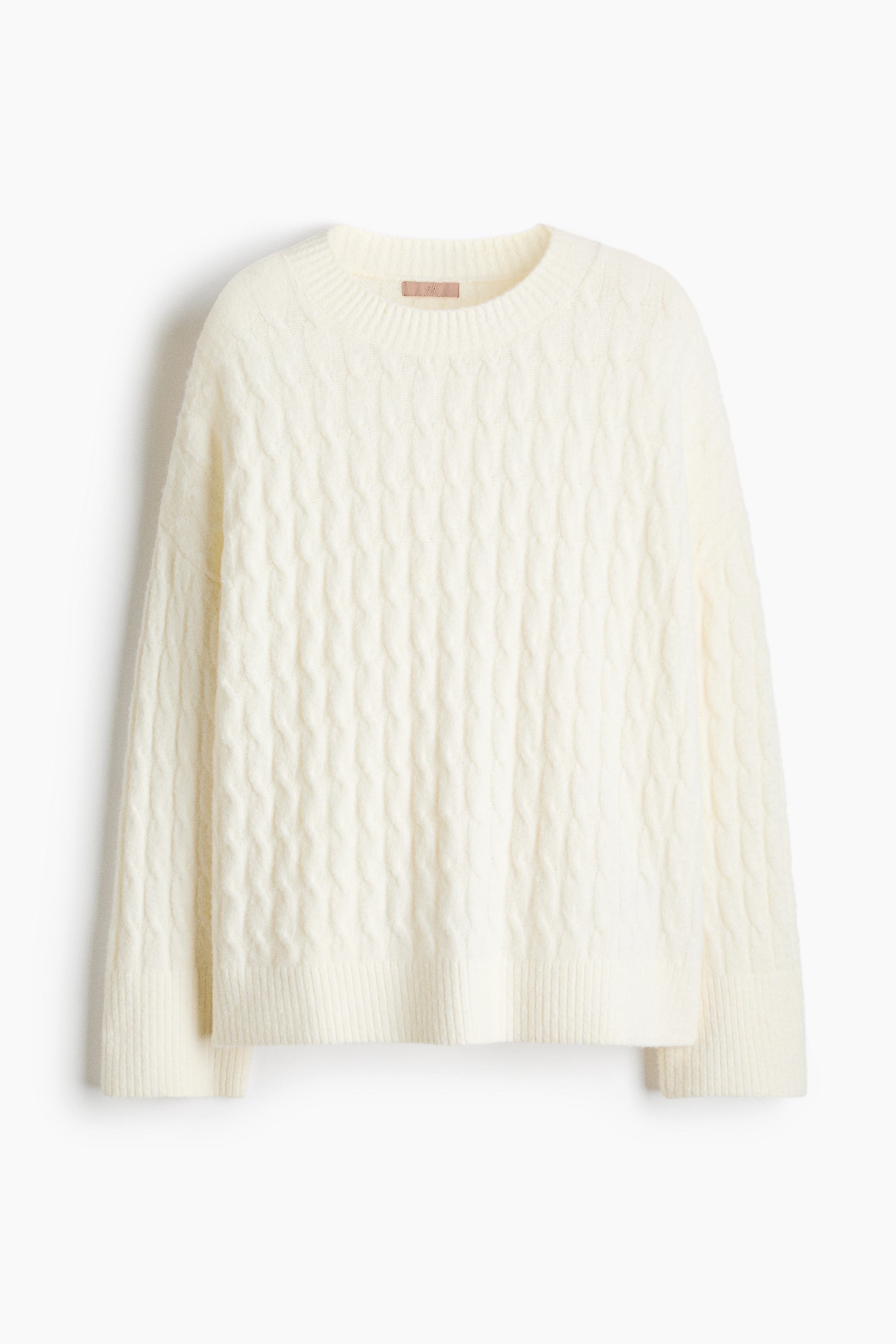 A light-coloured, loose-fit cable-knit jumper featuring a round neckline, dropped shoulders, and ribbed cuffs is being worn by the model, who is crossing their arms in front. The jumper is styled with dark, elastic-waist shorts.