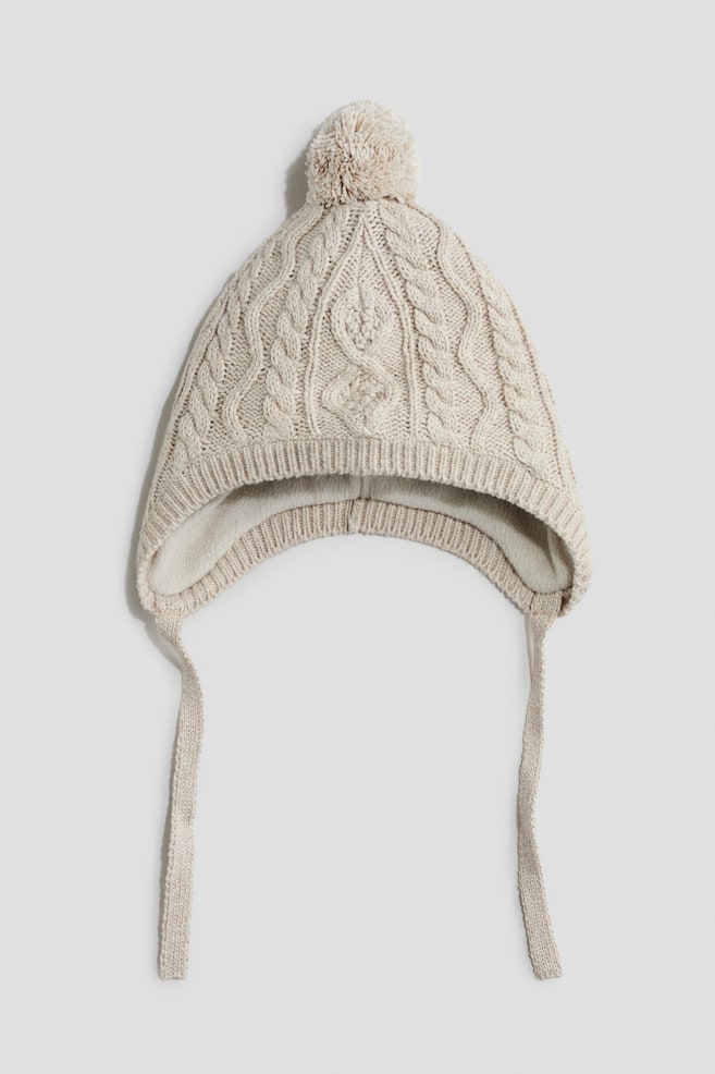 Eine cremefarbene StrickmÃ¼tze ist von vorne zu sehen und verfÃ¼gt Ã¼ber ein strukturiertes Zopfmuster, einen passenden Bommel oben sowie integrierte Ohrklappen mit langen KinnbÃ¤ndern, die nach unten hÃ¤ngen. Im Inneren ist ein weiches, flauschiges Fleecefutter sichtbar, das entlang des gerippten unteren Saums zu sehen ist. - 1