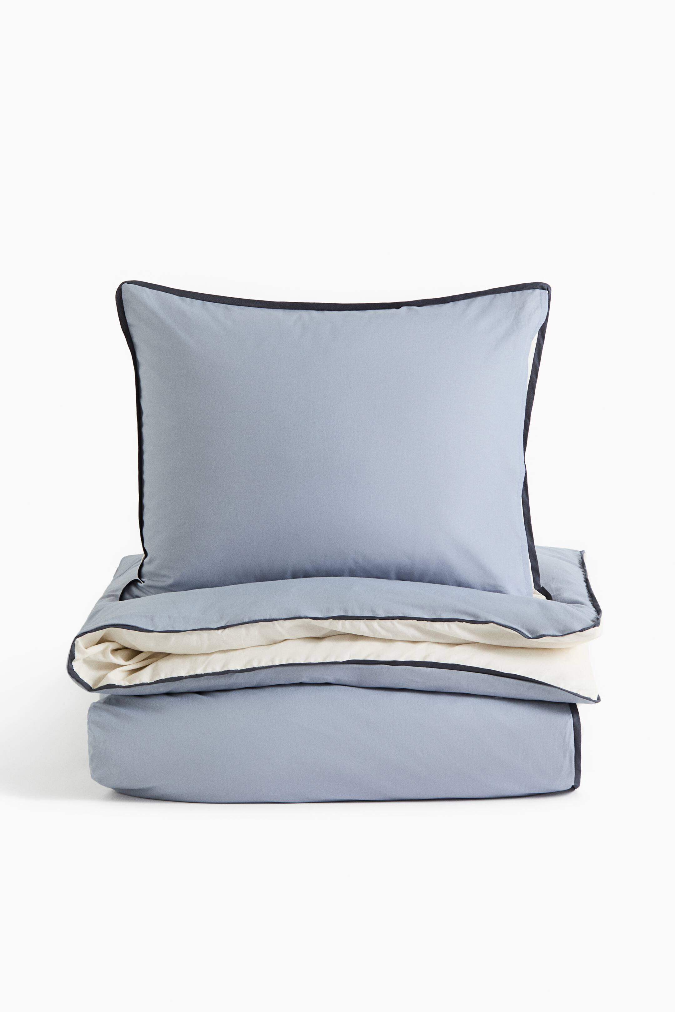 Agrandir l'image: Un ensemble de literie bleu clair présente un oreiller carré dressé à la verticale derrière une housse de couette pliée, orientée vers l’avant. Les deux pièces sont agrémentées d’un passepoil foncé contrastant le long des bords, tandis que la housse de couette pliée révèle un envers de couleur crème.
