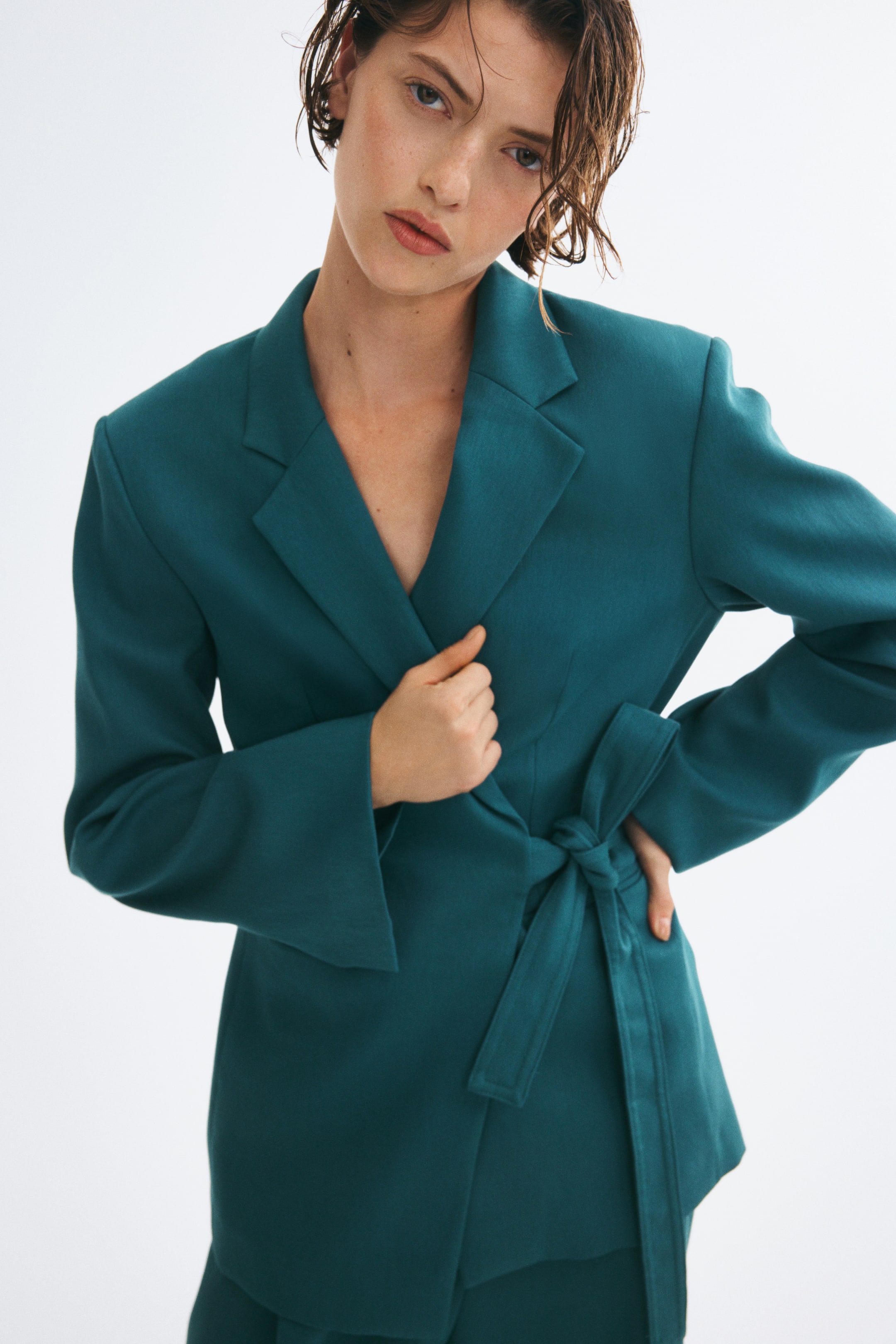 Zobraziť väčší obrázok: Model má na sebe oversized sako v tyrkysovej farbe s klopami do zárezu, prekladaným predným dielom a širokým zodpovedajúcim opaskom uviazaným v páse. Jednou rukou drží jednu z klop a druhú má položenú na boku.