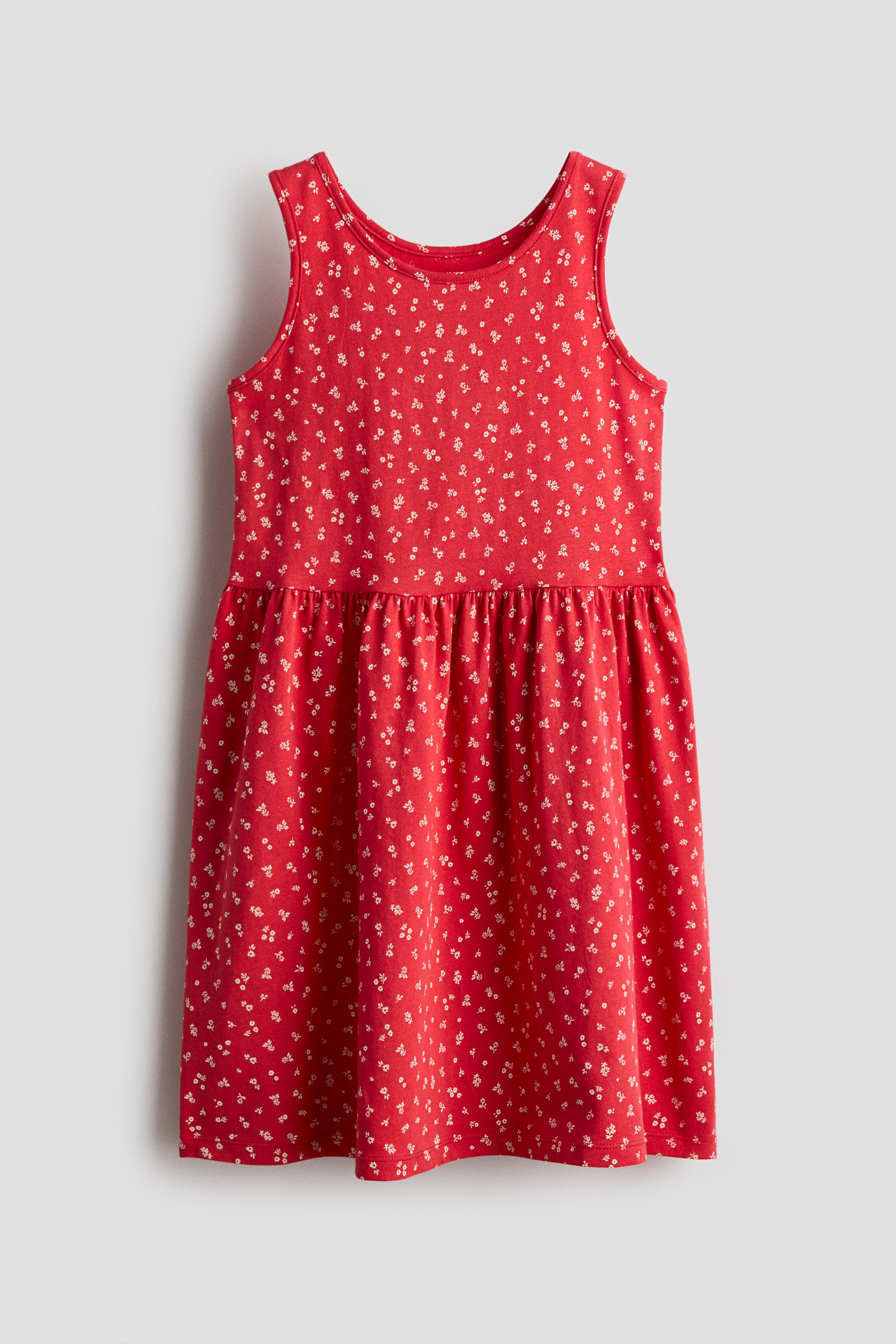 View larger image: A sleeveless red dress in soft cotton jersey is shown from the front, featuring an all-over white floral print and a round neckline. It shows a gathered seam at the waist leading to a flared skirt.