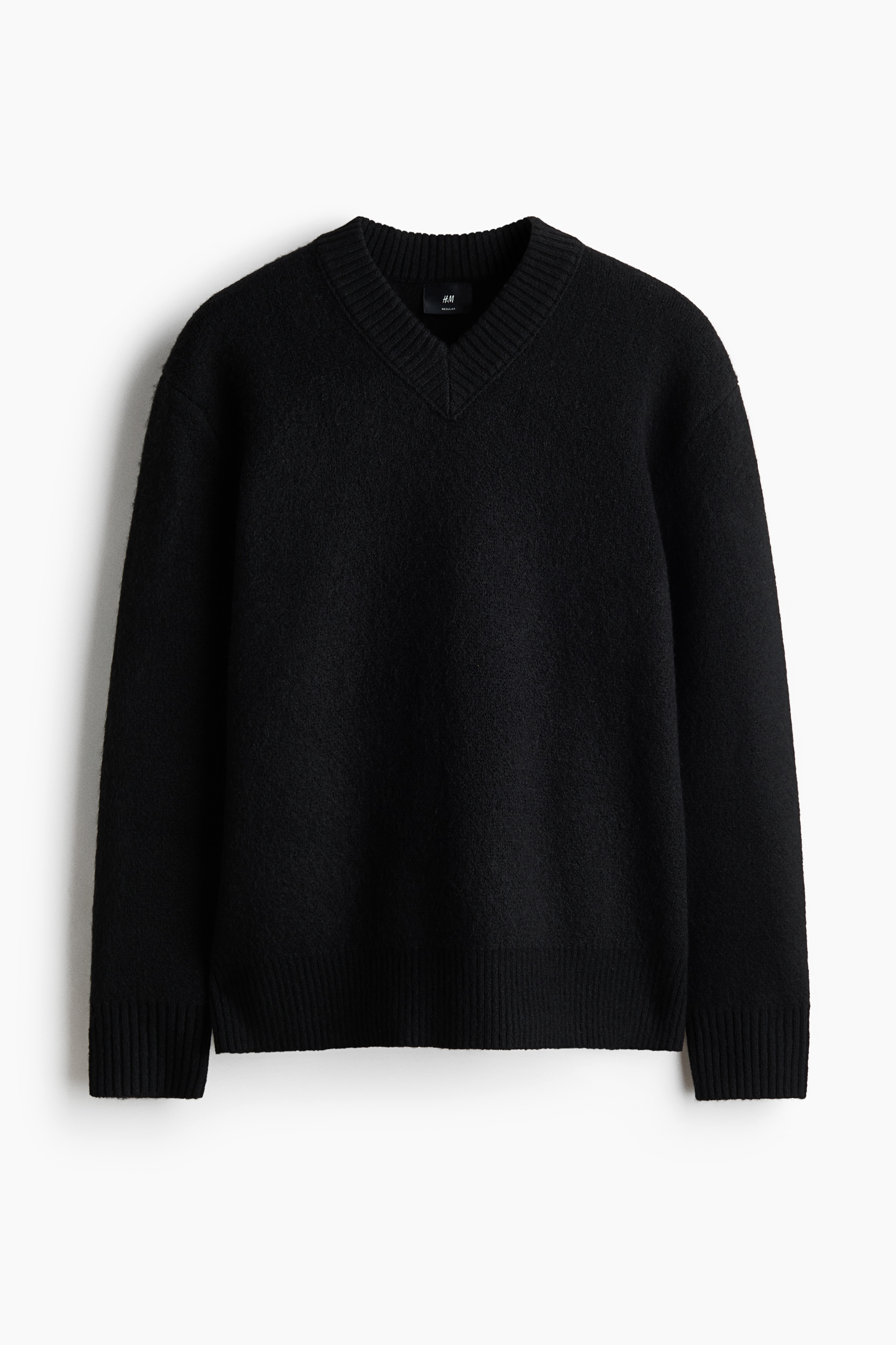 A black knit sweater featuring a relaxed fit, a ribbed V-neck, and ribbed trim at the cuffs and hem is being worn by the model. The sweater is styled with blue jeans and black sneakers.