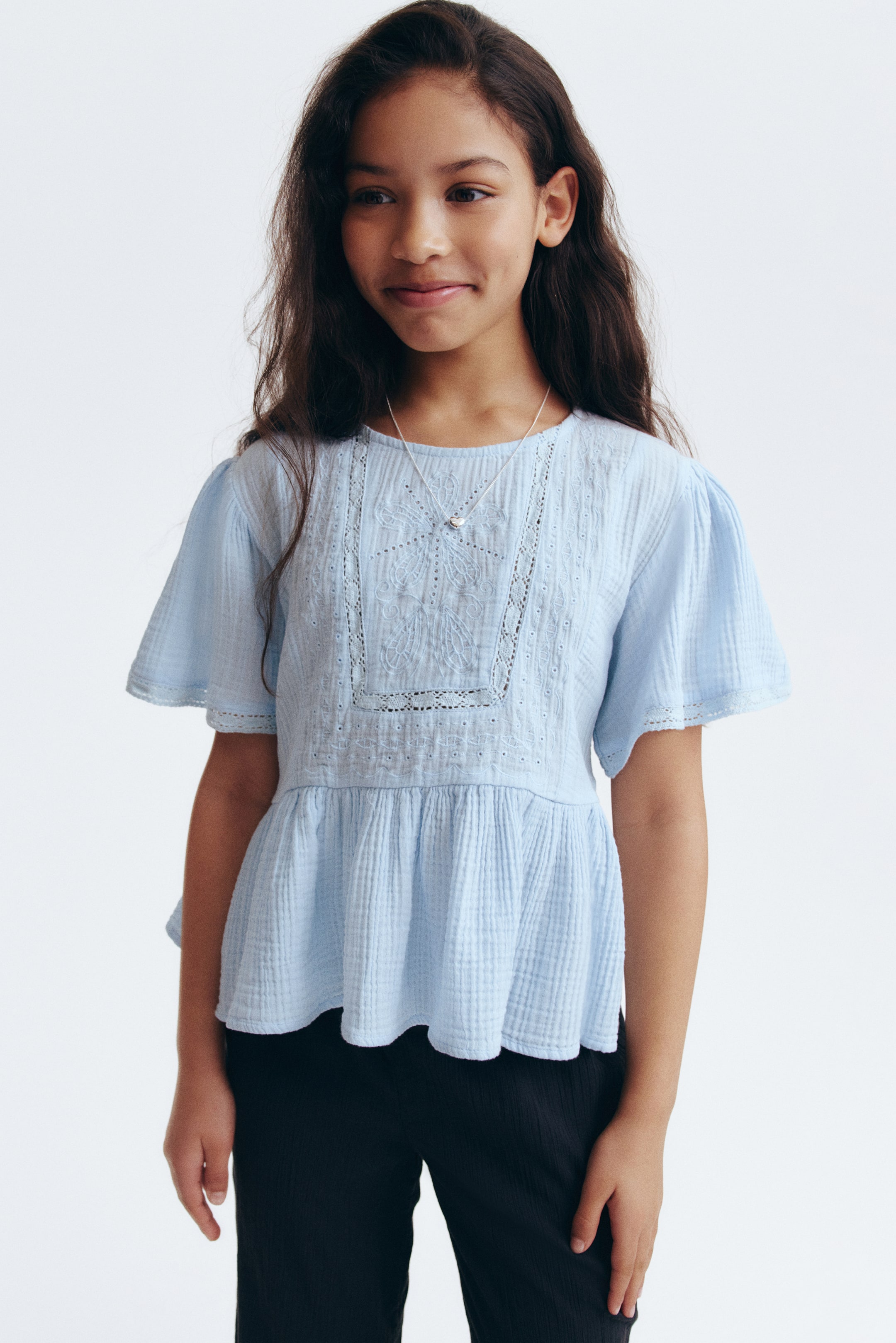 View larger image: The front of the light blue top is crafted from airy cotton muslin, showcasing broderie anglaise and lace insets. It features a round neckline and short, wide sleeves with gathers on the shoulders and lace-trimmed cuffs. The design includes a gathered seam at the waist, leading to a flared peplum.