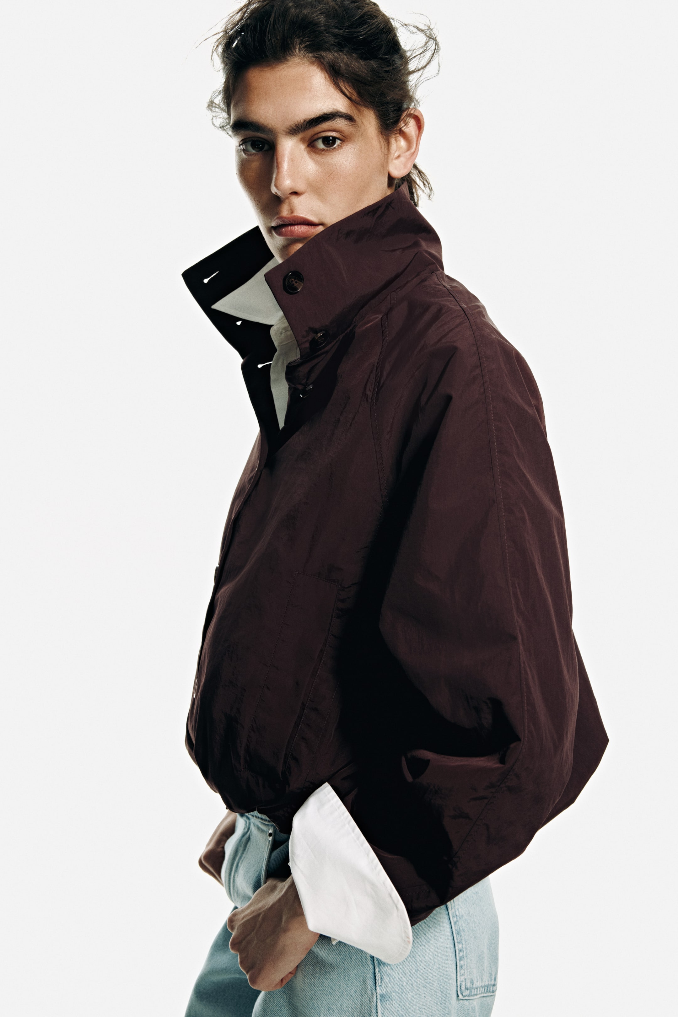 View larger image: The model is showcasing an oversized dark brown woven jacket characterized by a standing funnel collar with buttons and a buttoned front. It features long raglan sleeves with narrow elasticated cuffs and a gathered hem that forms a bubble silhouette. The jacket is layered over a white shirt, with light blue jeans visible at the waist.