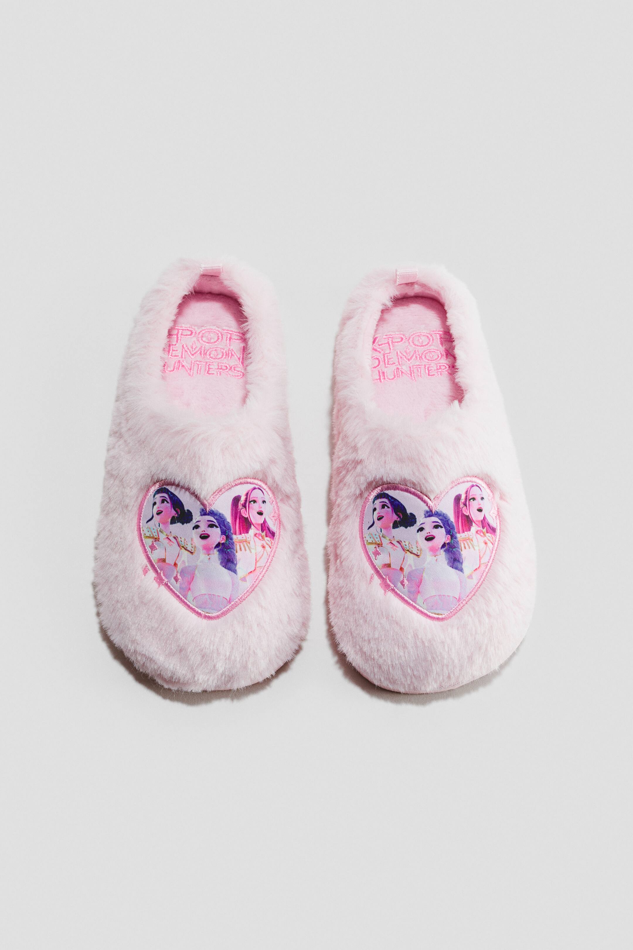 Agrandir l'image: Une paire de chaussons en fausse fourrure rose clair est présentée de face. Chaque chausson est ornée d’un patch en forme de cœur sur le dessus, représentant l’illustration de trois personnages féminins animés, tandis que la semelle intérieure rose laisse apparaître le texte « POP DEMON HUNTERS ».