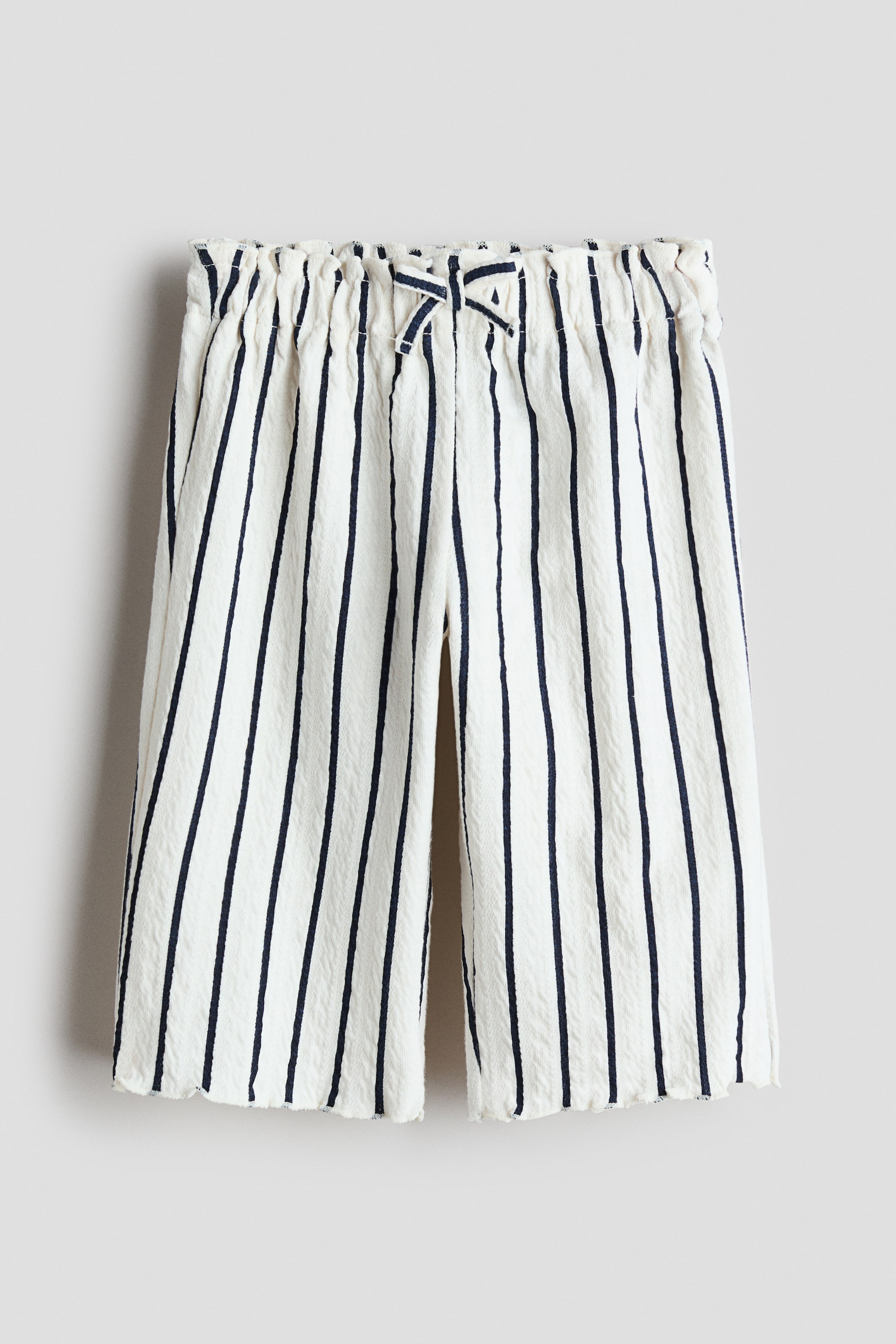 Ver imagen más grande: Los pantalones están confeccionados en un tejido de punto blanco texturizado con rayas verticales en azul marino, y presentan una cintura adornada con volantes y un pequeño lazo en la parte delantera. Ofrecen un ajuste holgado en la cadera y el muslo, con piernas anchas que terminan en dobladillos rematados.