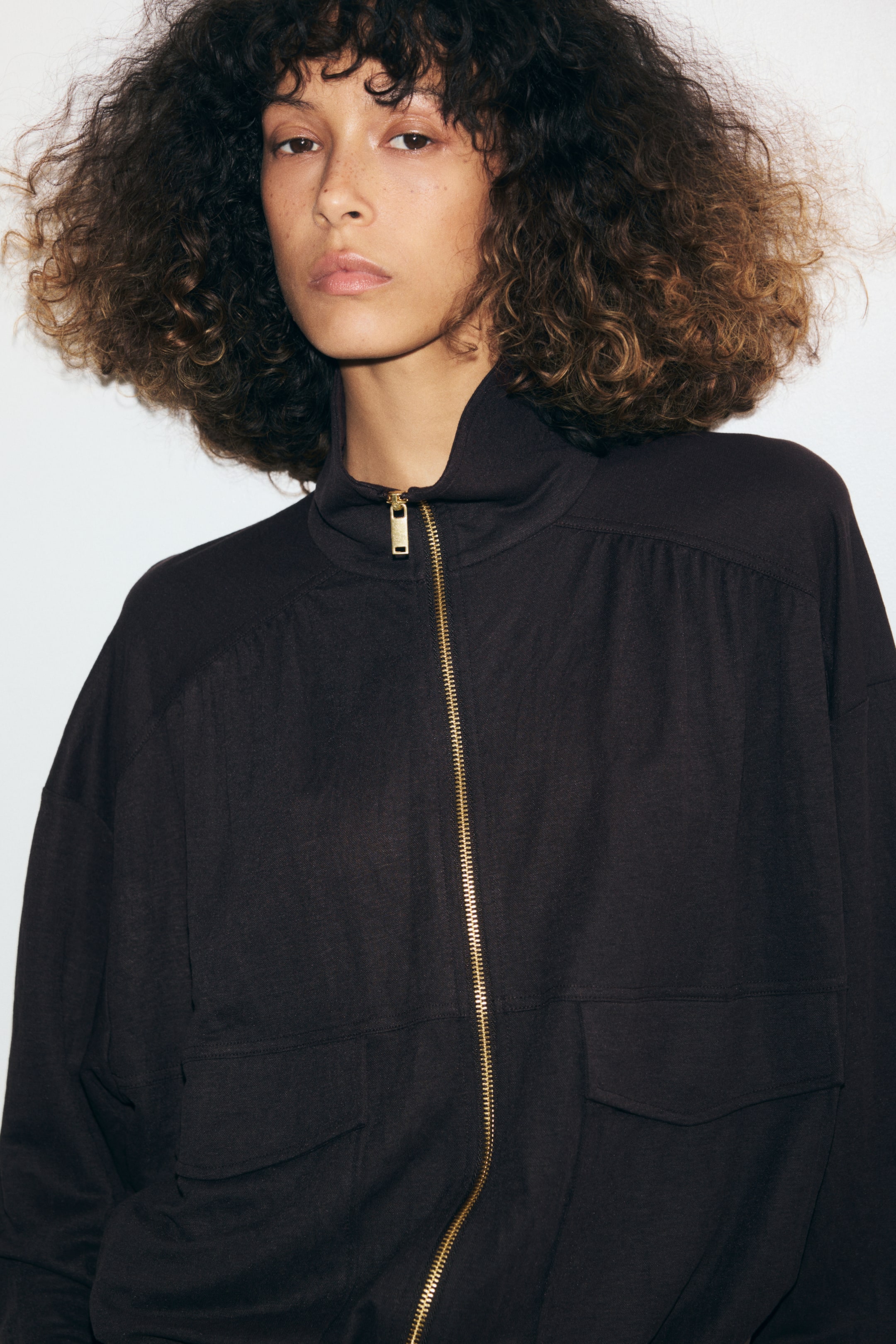 View larger image: A dark brown relaxed-fit jacket in a lightly crinkled fabric is worn by the model. The jacket features a high funnel collar, a gold-tone front zipper, dropped shoulders with gathered yoke detailing, and front patch pockets with flaps.