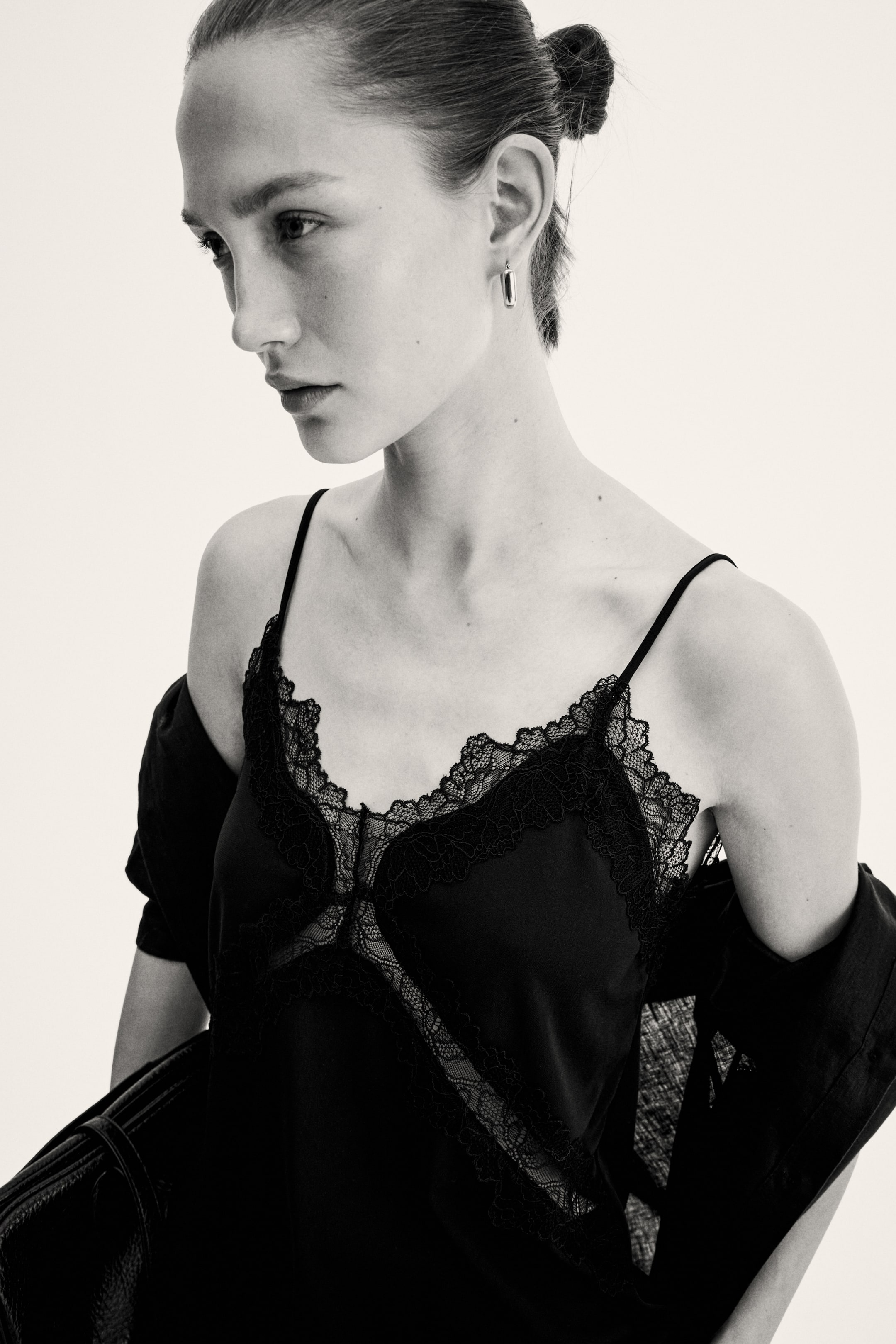 View larger image: A black camisole showcasing a scalloped lace trim along its V-neckline and a central lace panel, featuring thin spaghetti straps. It is viewed from a front-left angle, with a black jacket draped over the shoulders.