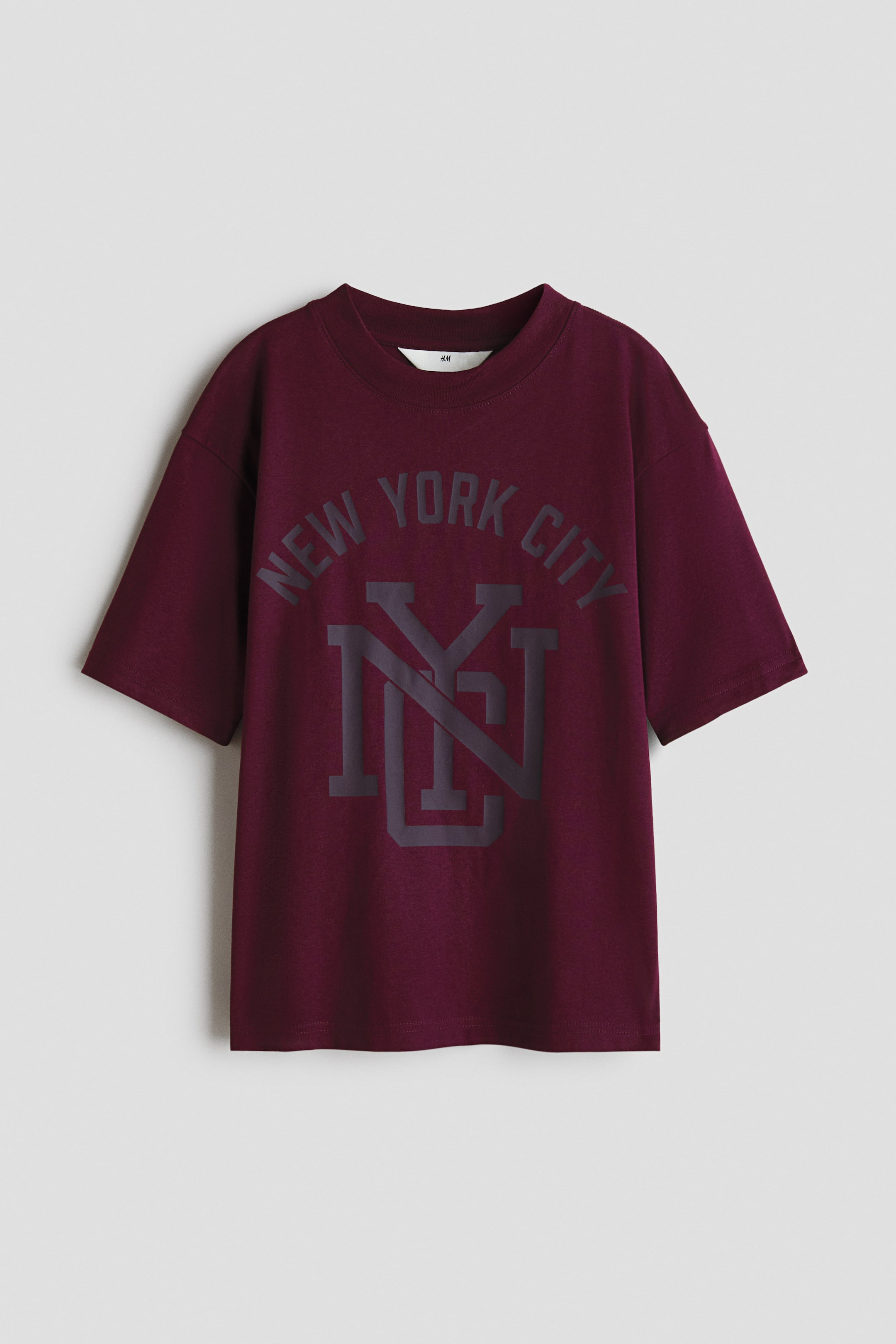 View larger image: Ang loose-fit na T-shirt ay ipinapakita mula sa harapan. Ito ay gawa sa maroon na cotton jersey at may ribbed trim sa paligid ng neckline at dropped shoulders. Isang dark gray na print ng "NEW YORK CITY" ang naka-arko sa ibabaw ng malaking, overlapping na "NY" graphic sa dibdib.