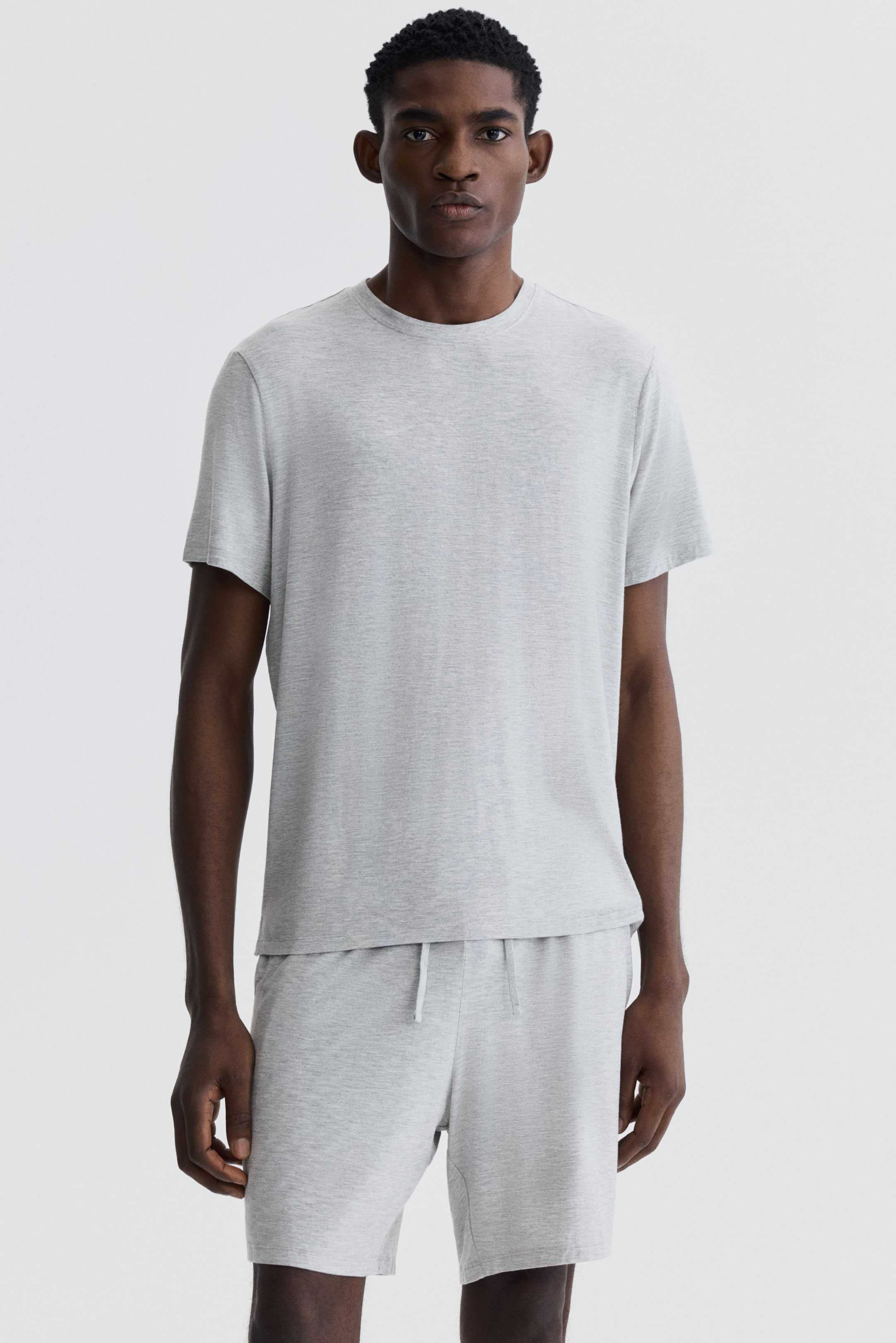 Ampliar la imagen: Se lleva puesta una camiseta de cuello redondo en color gris claro jaspeado, con mangas cortas y un ajuste relajado. También se usan shorts a juego en gris claro jaspeado, con cintura de cordón y que llegan por encima de la rodilla.