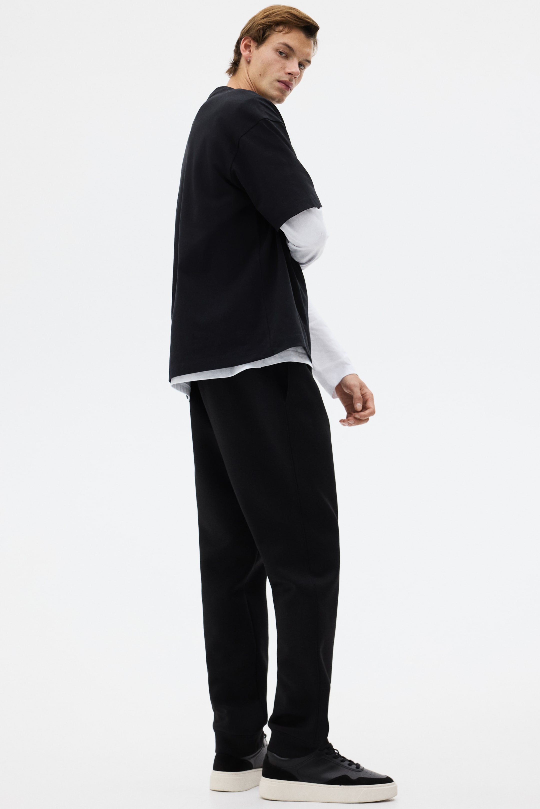 Größeres Bild anzeigen: Ein schwarzes übergroßes T-Shirt wird getragen, geschichtet über einem weißen Langarmshirt mit sichtbaren Manschetten und Saum, und wird von einem Dreiviertel-Rückenwinkel aus präsentiert, was seine lockere Passform hervorhebt. Schwarze Hosen mit lockerer Passform, die Seitentaschen und ein gerades Bein aufweisen, sind in dieser Ansicht ebenfalls sichtbar.