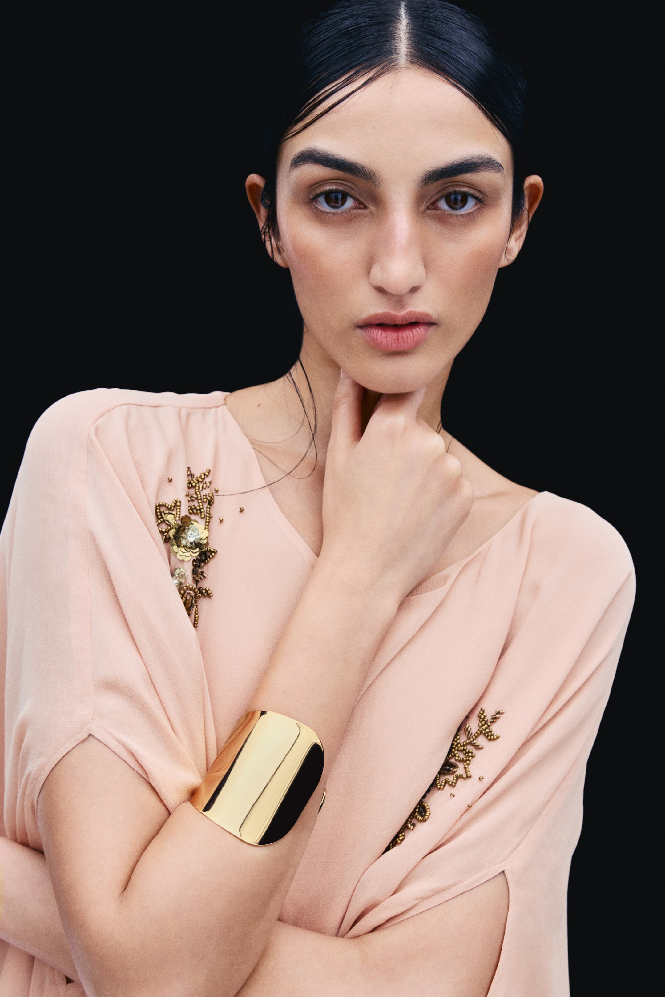 View larger image: A wide, shiny gold-toned cuff bracelet with an asymmetrical design is worn on the model’s forearm. The model rests their hand under their chin, highlighting the polished, reflective finish of the jewellery.