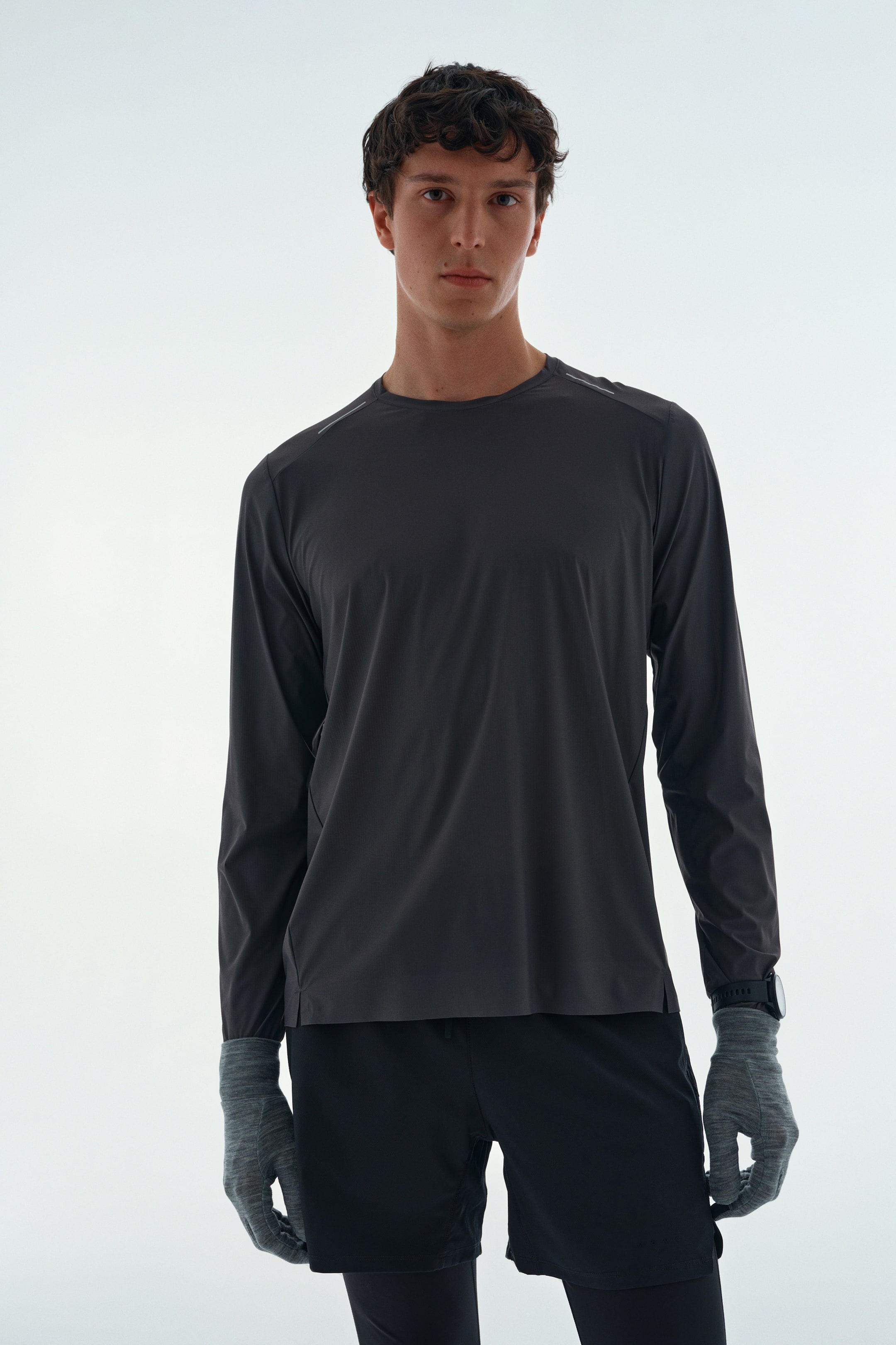 View larger image: A dark grey long-sleeve athletic top is worn by the model, featuring a crew neck and subtle reflective details on the shoulders. The top is styled with black athletic shorts, dark tights, and grey gloves.