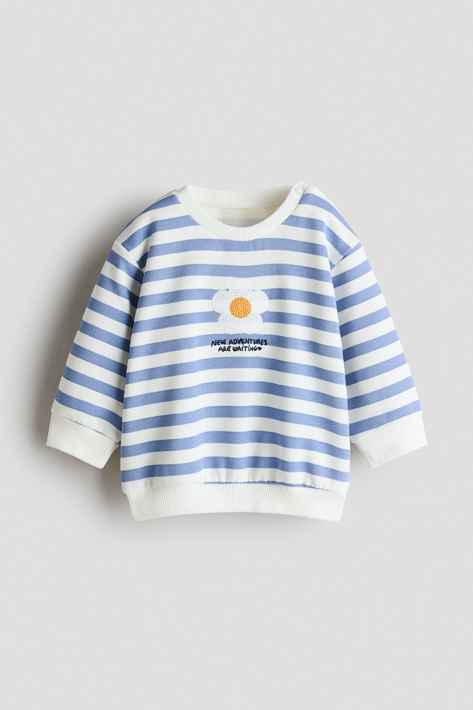 En langÃ¦rmet sweatshirt set forfra med blÃ¥ og hvide vandrette striber, komplet med en ribstrikket rund hals, ribstrikkede manchetter og en ribstrikket kant. Midt pÃ¥ brystet ses et struktureret hvidt blomsterpatch med gul midte, placeret over den broderede sorte tekst "NEW ADVENTURES ARE WAITING".