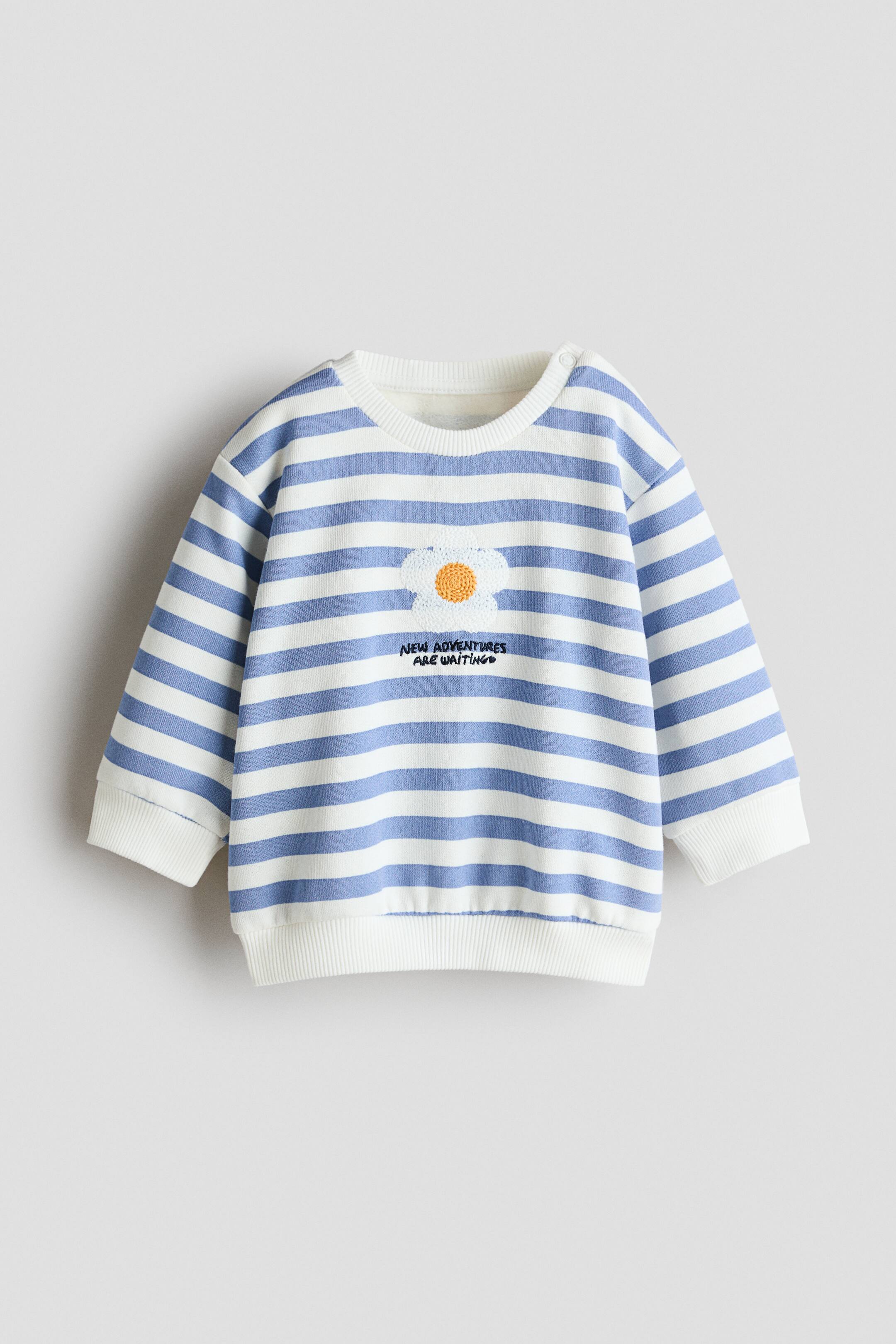 Se større billede: En langærmet sweatshirt set forfra med blå og hvide vandrette striber, komplet med en ribstrikket rund hals, ribstrikkede manchetter og en ribstrikket kant. Midt på brystet ses et struktureret hvidt blomsterpatch med gul midte, placeret over den broderede sorte tekst "NEW ADVENTURES ARE WAITING".