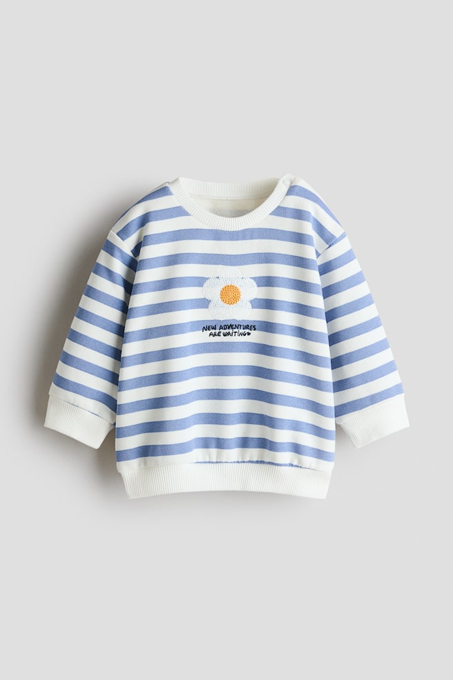 Ein frontseitig abgebildeter Langarm-Sweatshirt ist mit blauen und weiÃen horizontalen Streifen versehen und verfÃ¼gt Ã¼ber einen gerippten Rundhalsausschnitt, gerippte BÃ¼ndchen sowie einen gerippten Saum. Auf der Brustmitte ist ein strukturiertes weiÃes Blumen-Patch mit gelber Mitte zu sehen, das sich oberhalb des aufgestickten schwarzen Schriftzugs âNEW ADVENTURES ARE WAITINGâ befindet. - 1