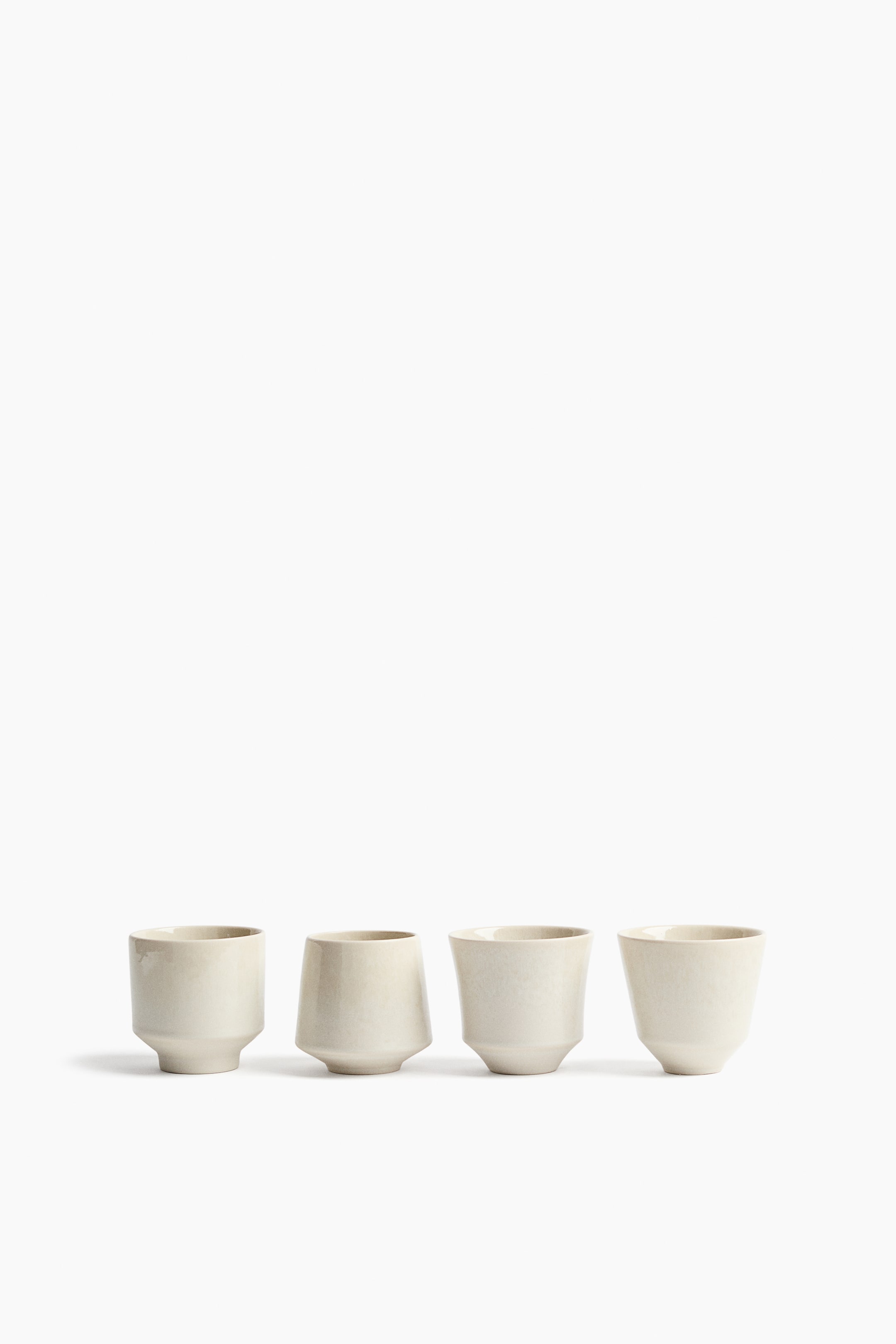 View larger image: Four cream-colored ceramic cups are arranged in a row, each featuring a distinct shape and a textured matte finish. The leftmost cup has a cylindrical upper body that tapers to a narrow base. The second cup shows an inverted trapezoidal shape with angled sides. The third cup exhibits a gentle S-curve profile, and the rightmost cup displays a tapered, inverted cone shape.