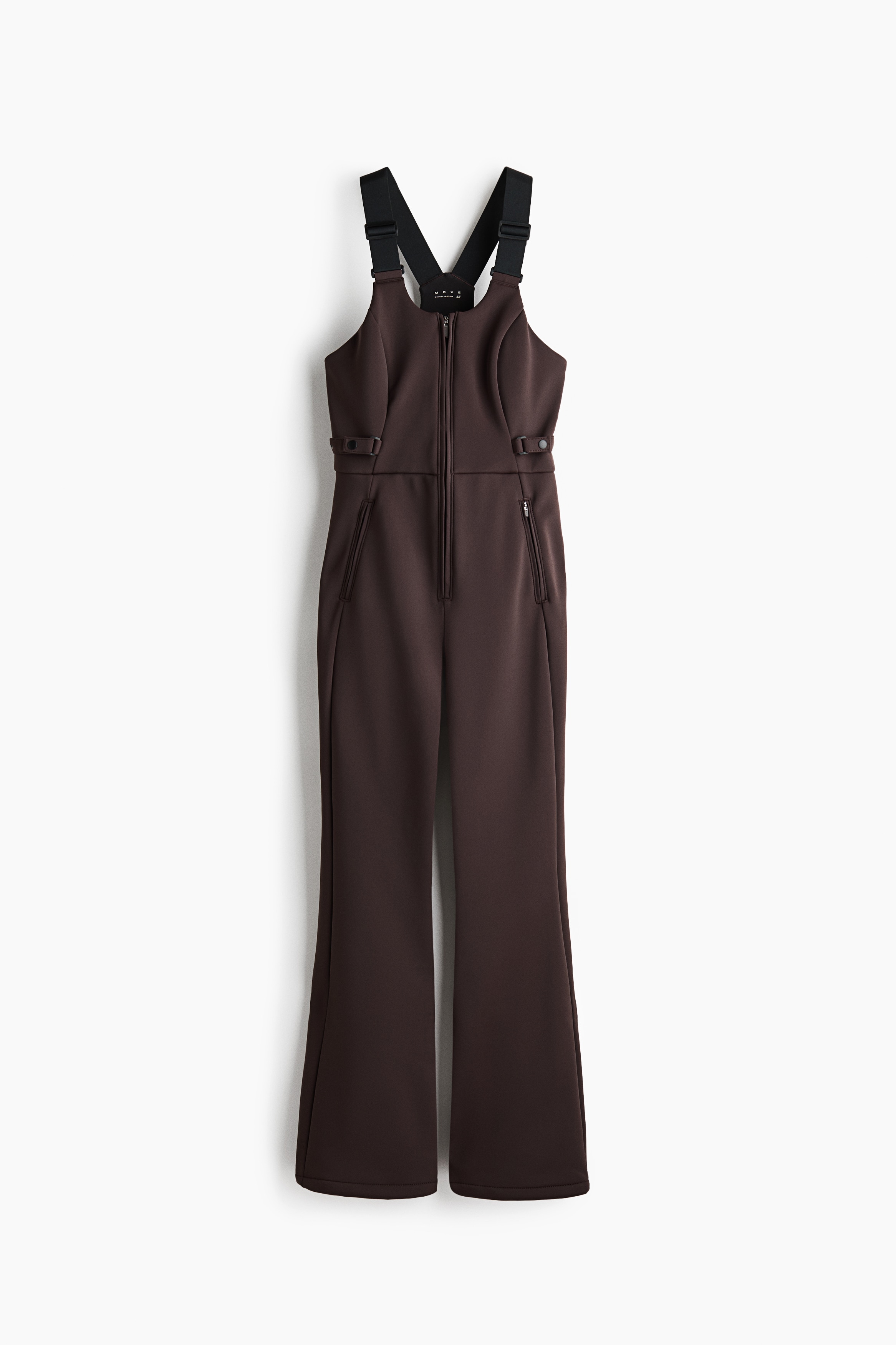 Dark brown ski overalls are being worn by the model, layered over a black long-sleeved turtleneck sweater. The slim-fitting overalls feature a high-waisted bib, side-button detailing, and bootcut legs that are resting over black ski boots.