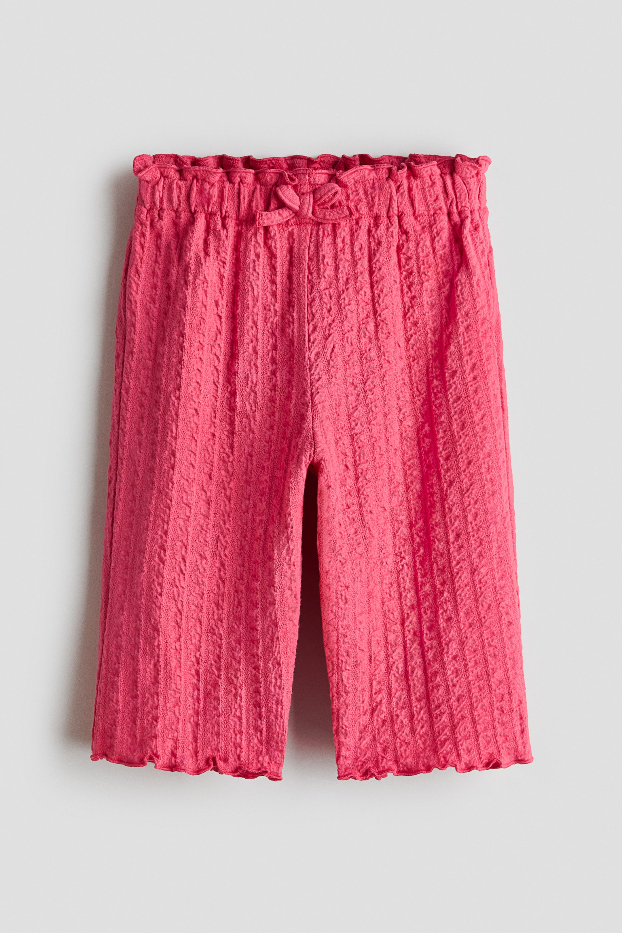 Ampliar la imagen: Los pantalones rosa de punto presentan una pretina elástica fruncida con un pequeño lazo decorativo en la parte frontal. Cuentan con rayas verticales texturizadas a lo largo de las piernas y un dobladillo con volantes y borde festoneado.
