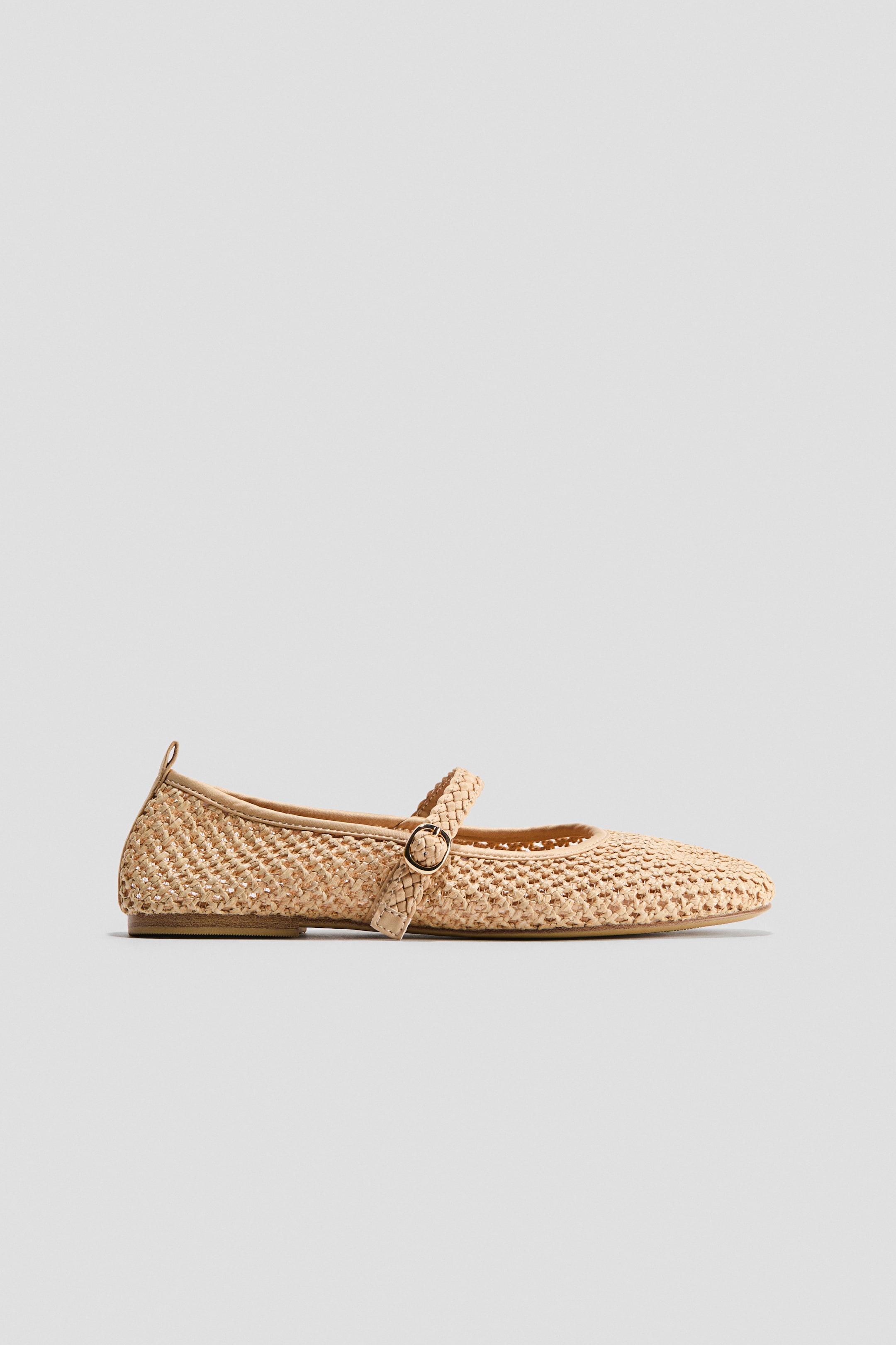 Ampliar la imagen: Un zapato plano estilo Mary Jane muestra su perfil lateral, confeccionado en rafia tejida de color beige claro natural con una punta afilada y una suela plana delgada. Presenta una correa trenzada con una hebilla redonda de tono dorado antiguo que se extiende a través del empeine.