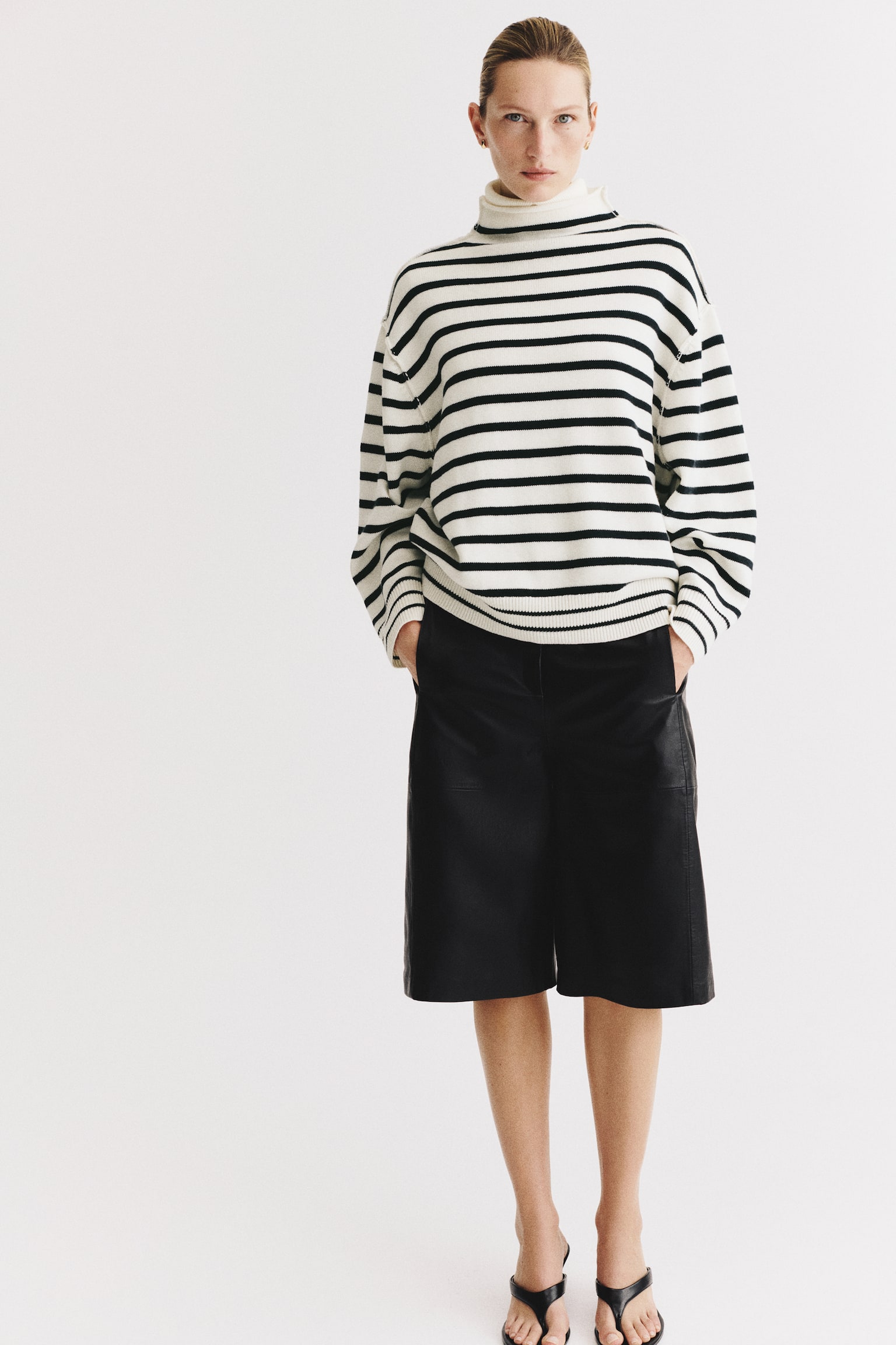 An oversized, cream-coloured knit sweater with horizontal black stripes is being worn by the model. The sweater features a high neck with a rolled edge, dropped shoulders, and ribbed trim at the cuffs and hem, paired with black knee-length shorts. - 1