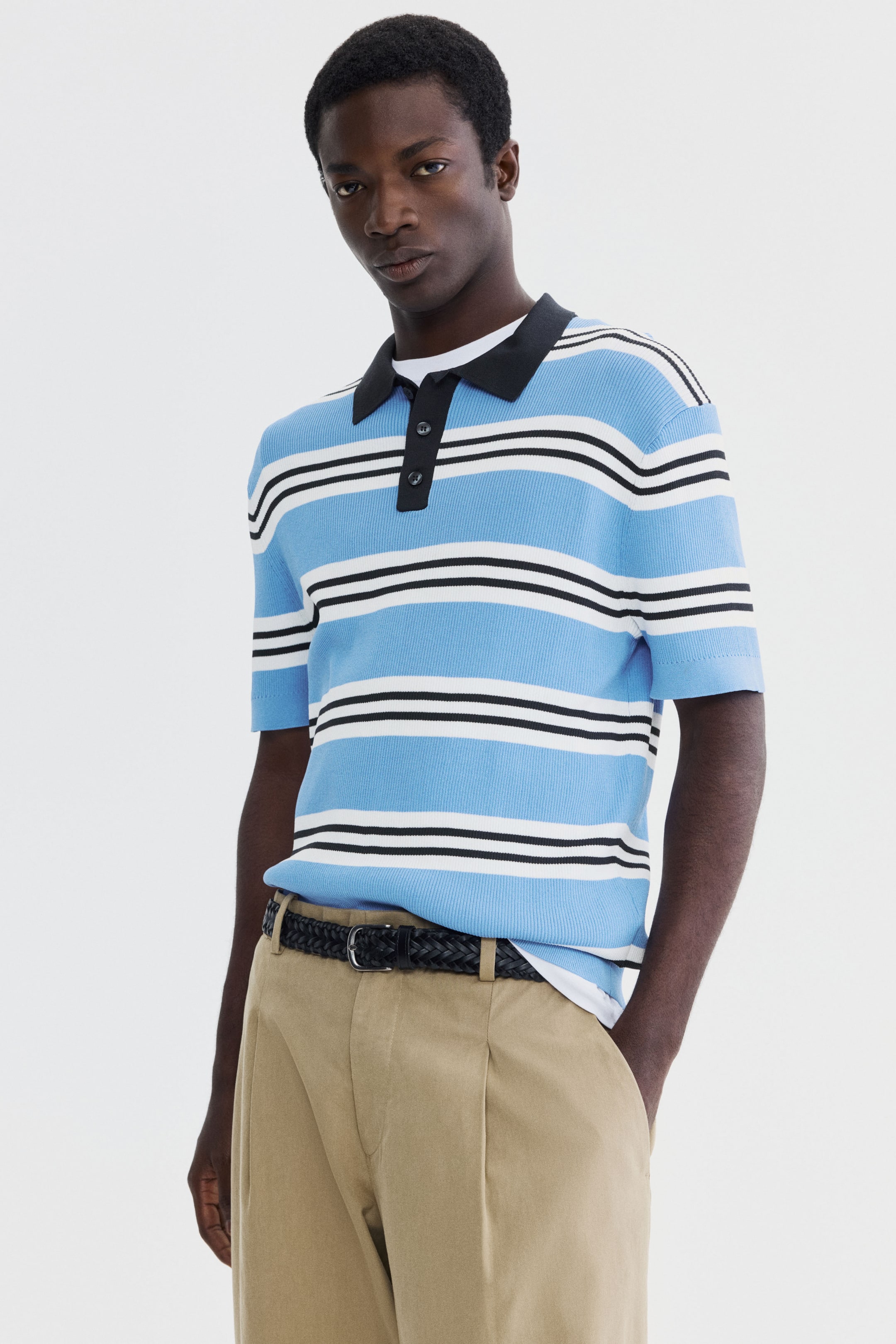 View larger image: The front-facing polo shirt showcases a light blue ribbed knit adorned with horizontal white and black stripes. Crafted with a black collar, a three-button black placket, and short sleeves, it subtly reveals a white undershirt beneath.